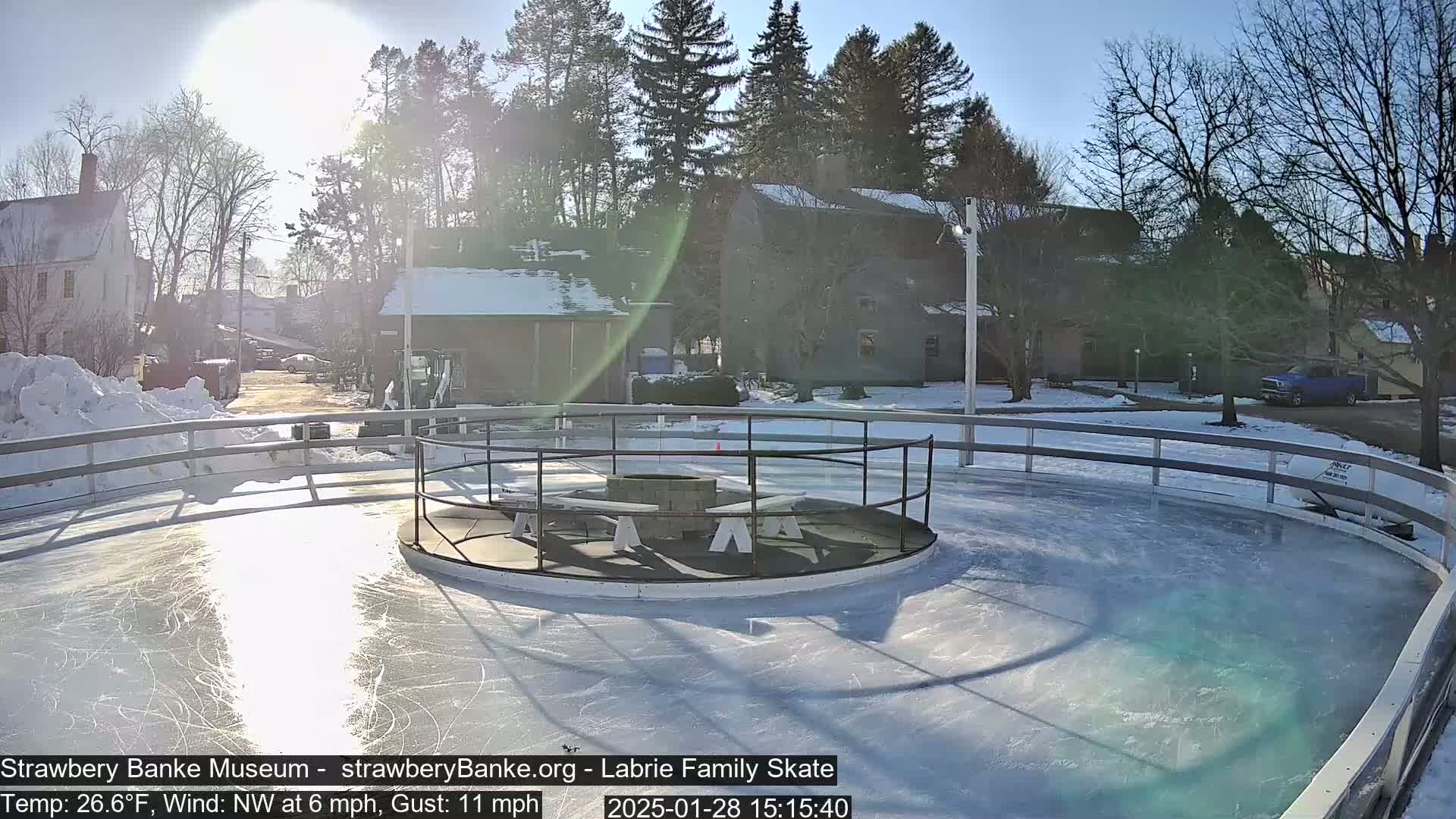 A sunny, partially snow-covered outdoor ice skating rink with a fire pit in the center, surrounded by buildings and trees.