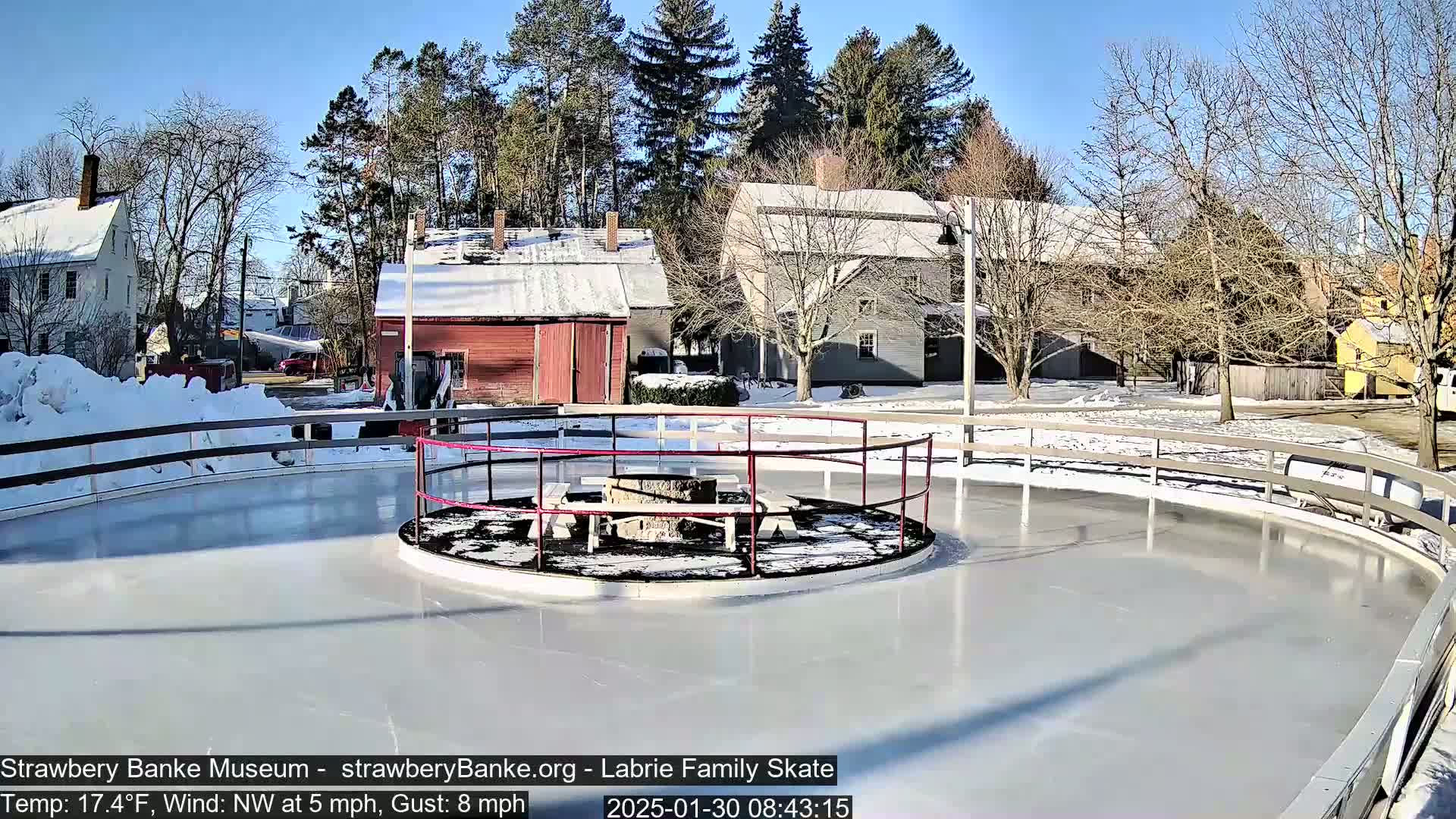 A sunny outdoor ice rink with a central fire pit and surrounding benches is nestled between snow-covered buildings and trees.