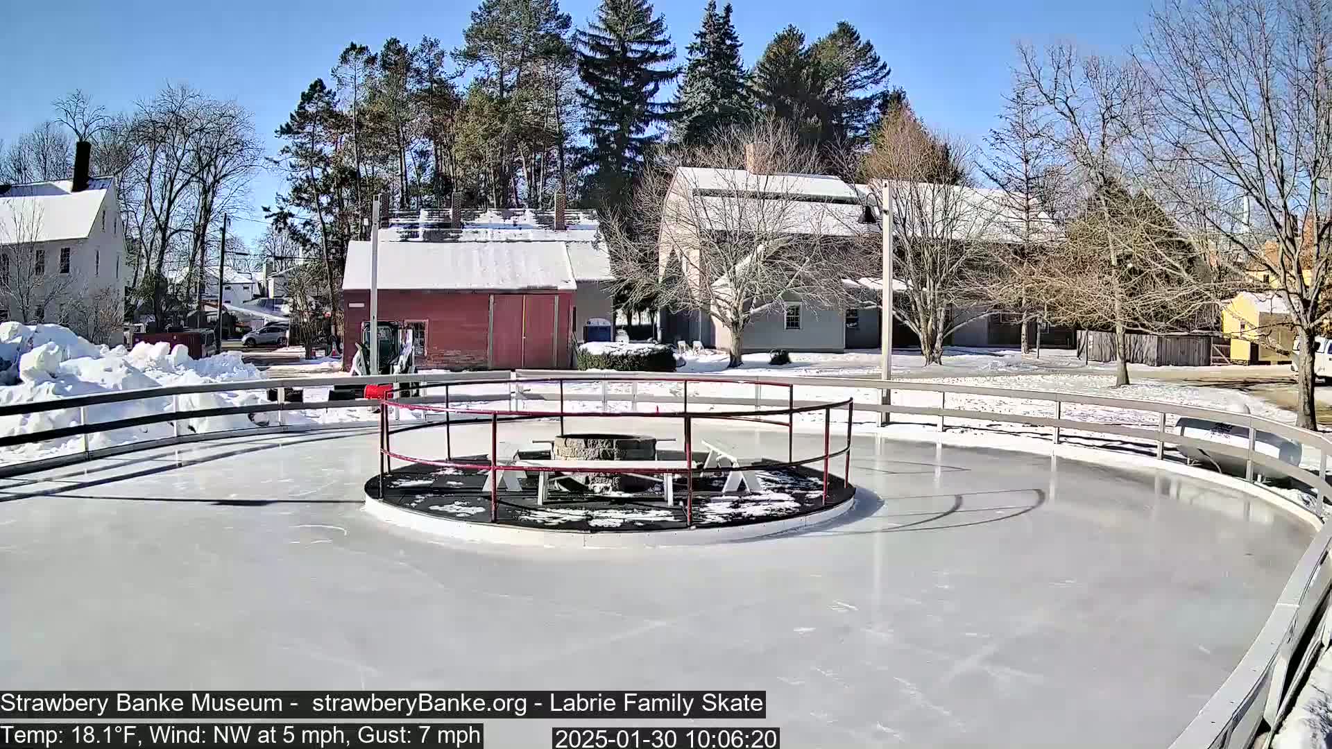 A round outdoor ice skating rink with a fire pit in the center, surrounded by snow-covered buildings and trees under a sunny sky.