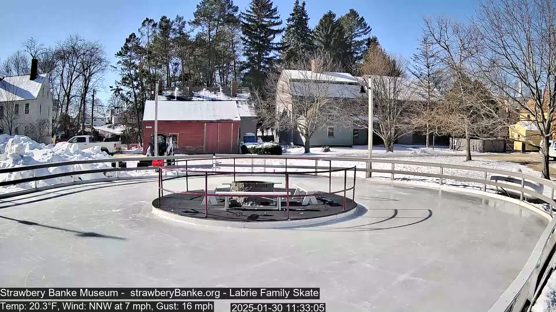 A sunny outdoor ice skating rink with a fire pit in the center, surrounded by snow-covered buildings and trees.
