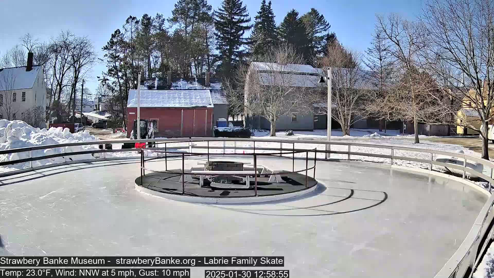 A circular ice rink with a fire pit and benches in the center is surrounded by a low fence, situated outdoors on a sunny day with snow on the ground and nearby buildings.