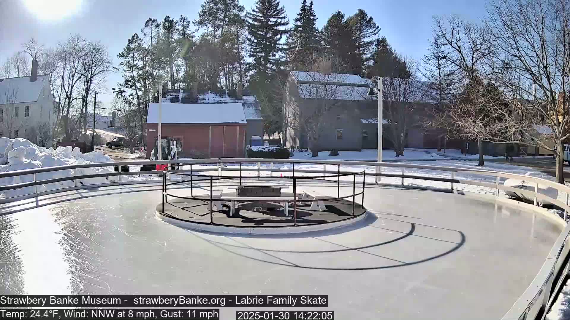 A sunny outdoor ice skating rink, surrounded by snow and buildings, shows some skating marks on its surface.