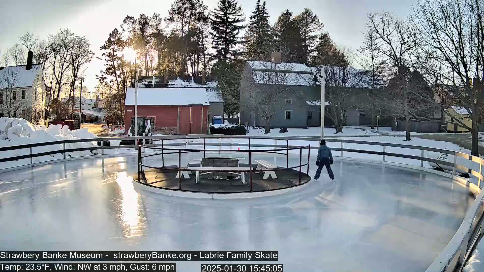 A person ice skates on an outdoor rink surrounded by snow-covered buildings and trees under a partly sunny sky.