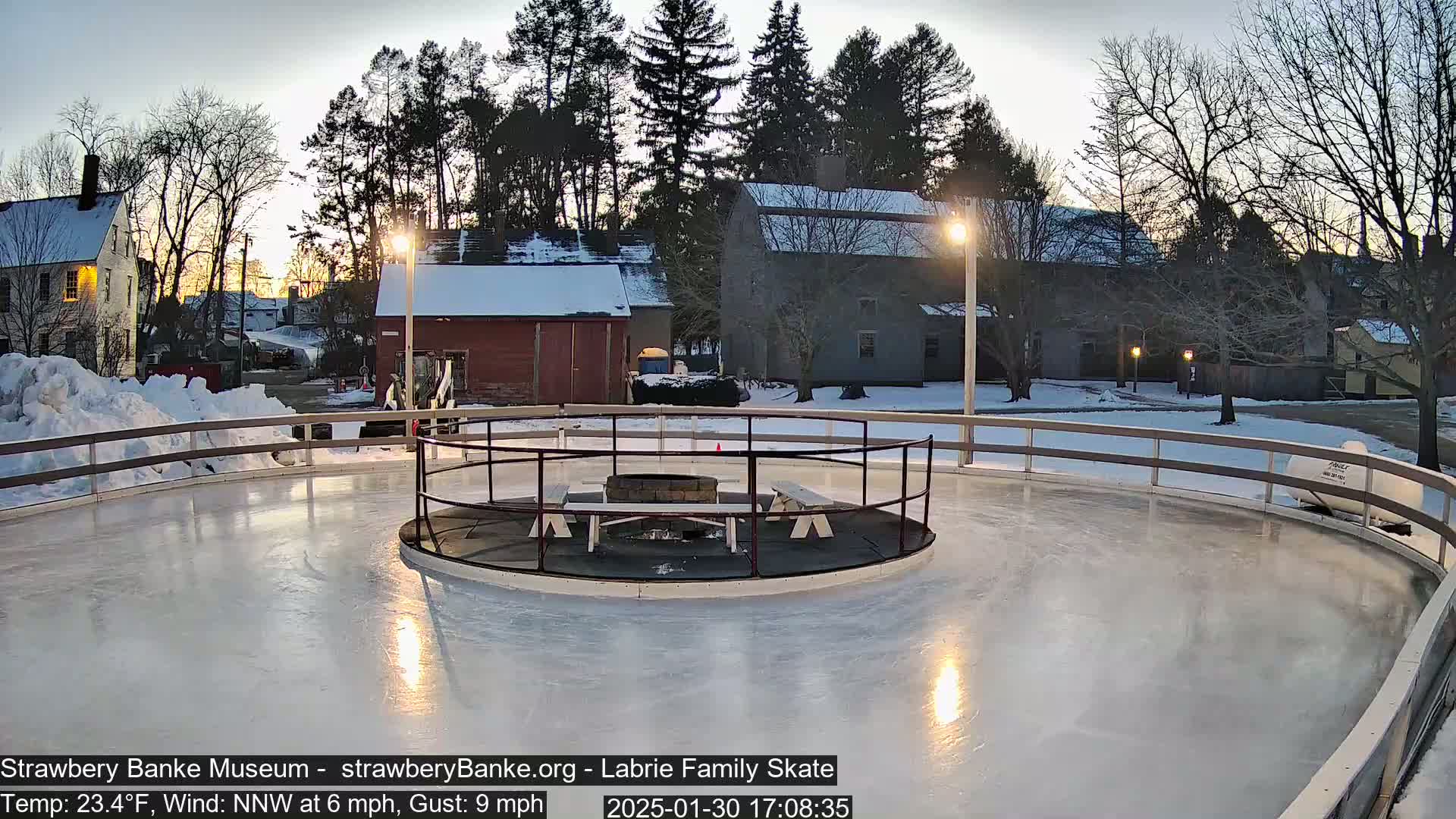 An outdoor ice skating rink with a central fire pit and benches is surrounded by snow-covered buildings and trees under a twilight sky.
