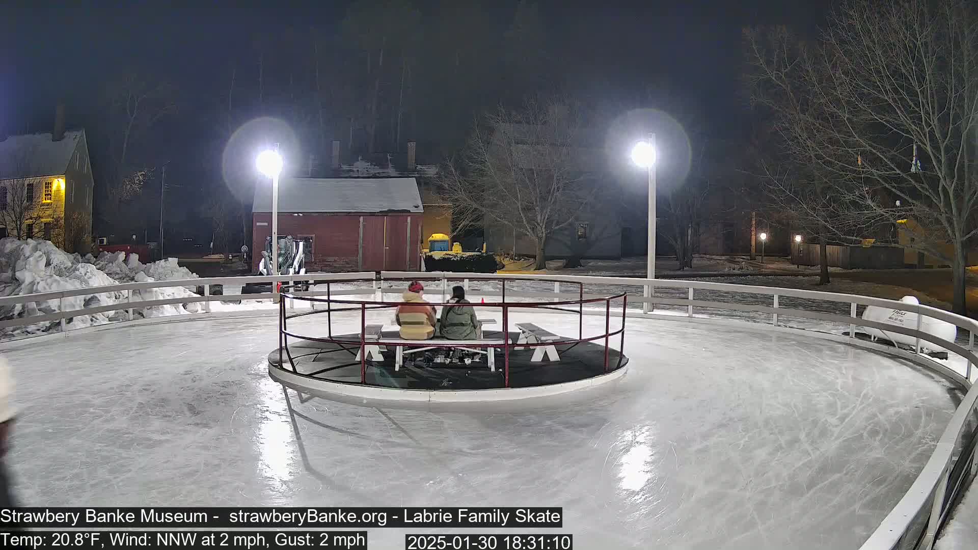 Two people sit on a bench in the center of an outdoor, illuminated ice rink at night, with a light snow cover on the surrounding ground.