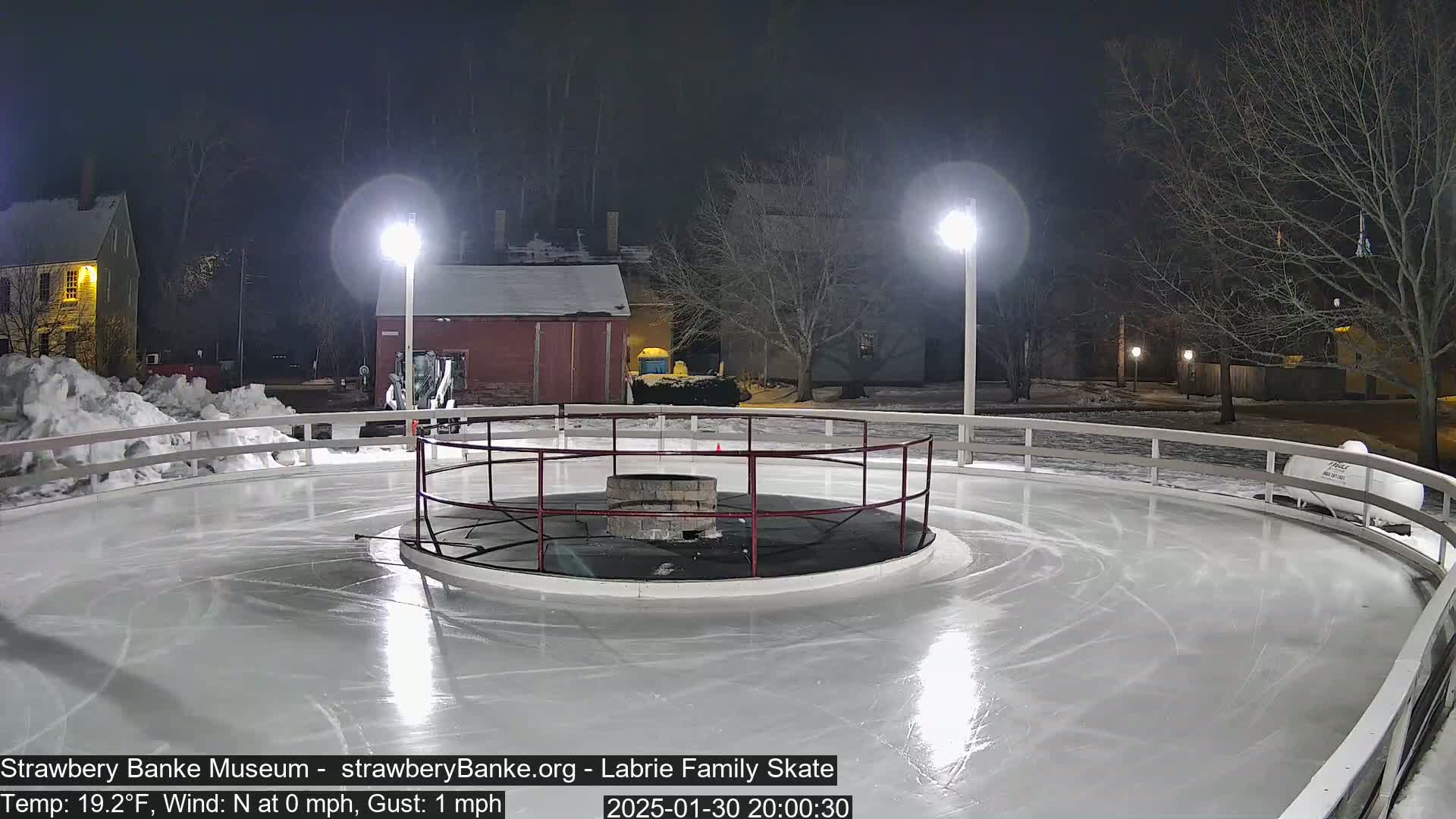 An outdoor ice skating rink at night, illuminated by lights, is surrounded by a low white fence with a central fire pit, and shows some skating marks on the ice.