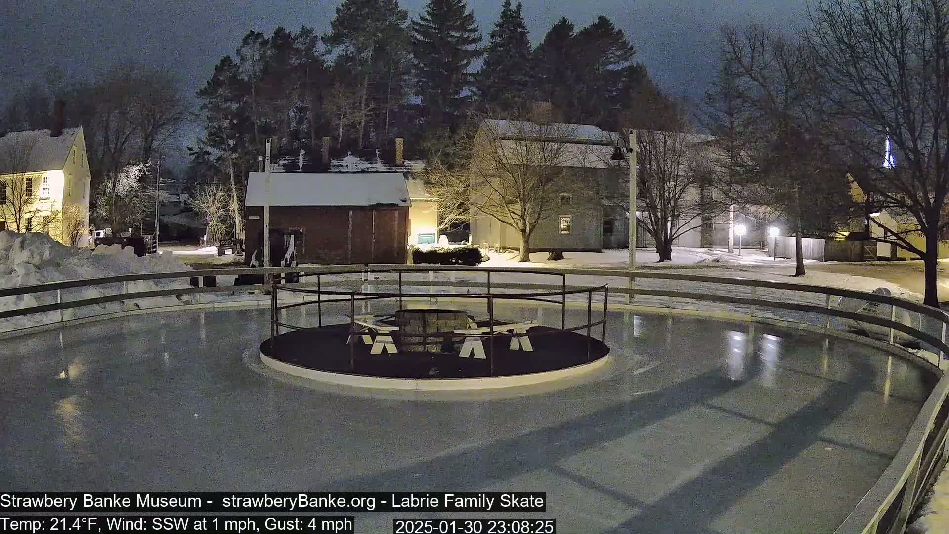 An outdoor ice rink, surrounded by snow-covered ground and buildings under a dark night sky, with a small fire pit in the center.