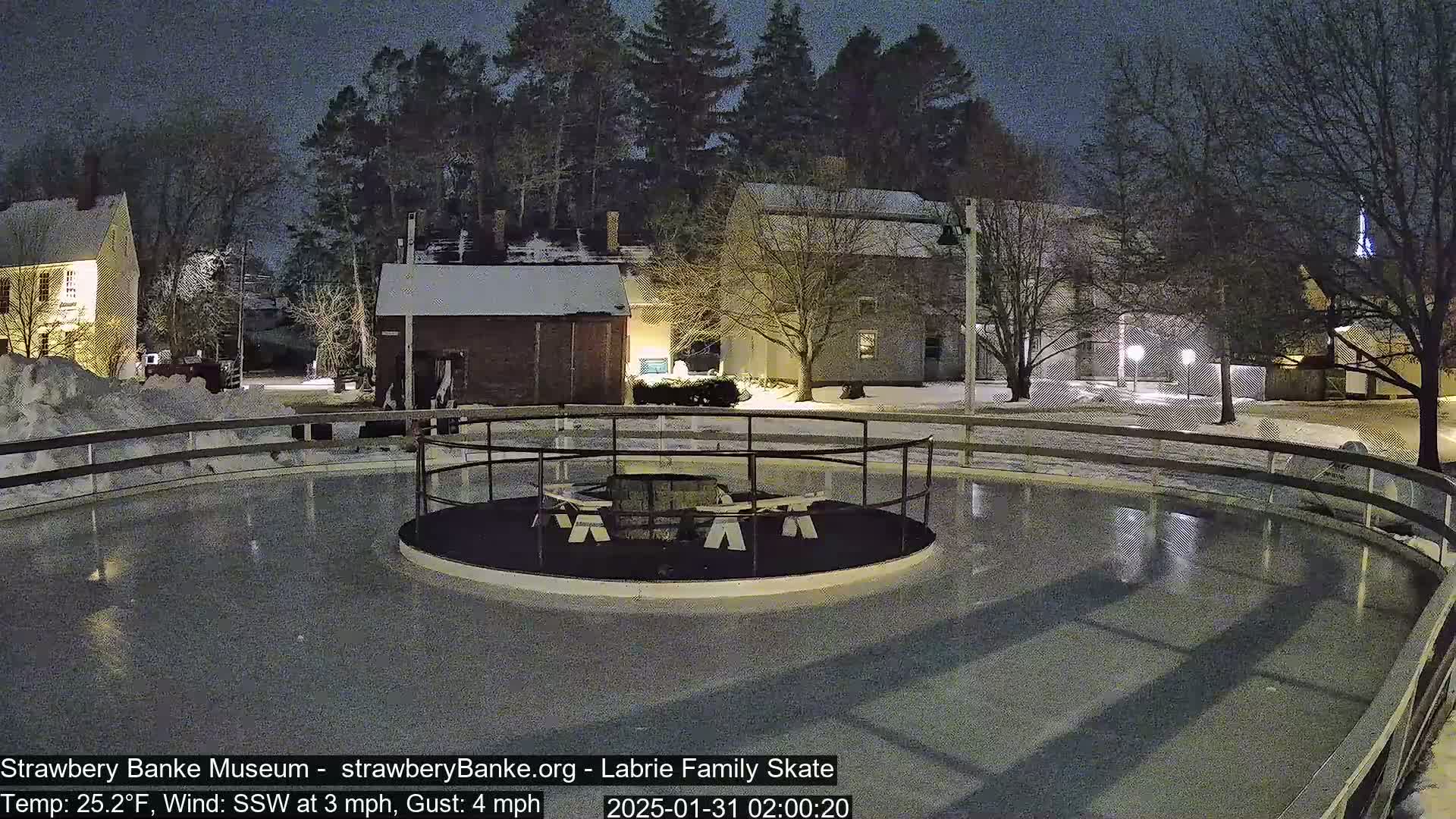 An outdoor ice skating rink, lit at night, is surrounded by snow-covered ground and buildings under a dark sky.