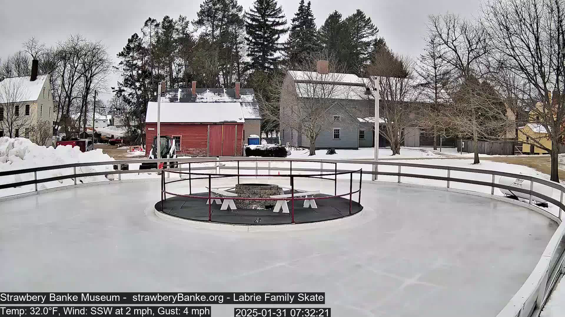 A snow-covered outdoor ice skating rink with a central fire pit and benches, surrounded by buildings and trees under an overcast sky.