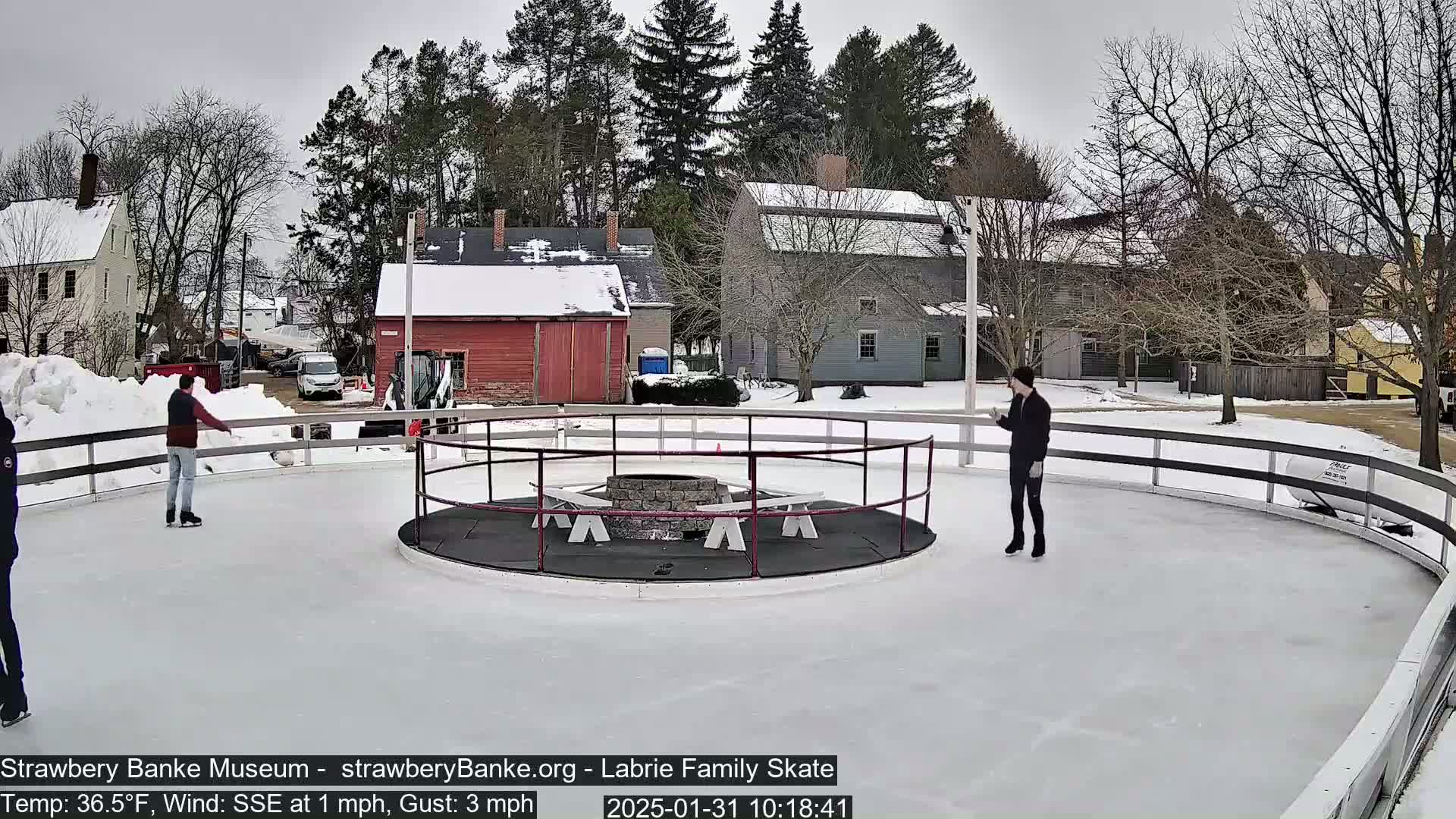 Three people ice skate on an outdoor rink surrounded by a snow-covered landscape and historic buildings, under a cloudy sky with a southeasterly wind.
