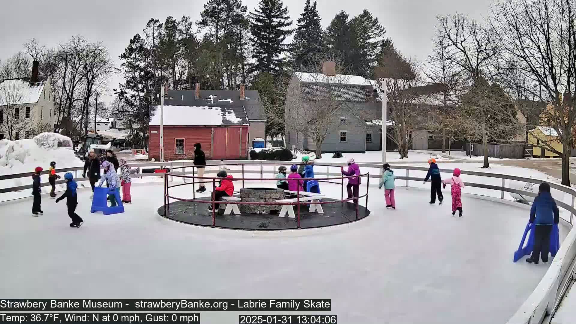 Children ice skate on an outdoor rink surrounded by a snow-covered landscape and historic buildings on a cloudy day.