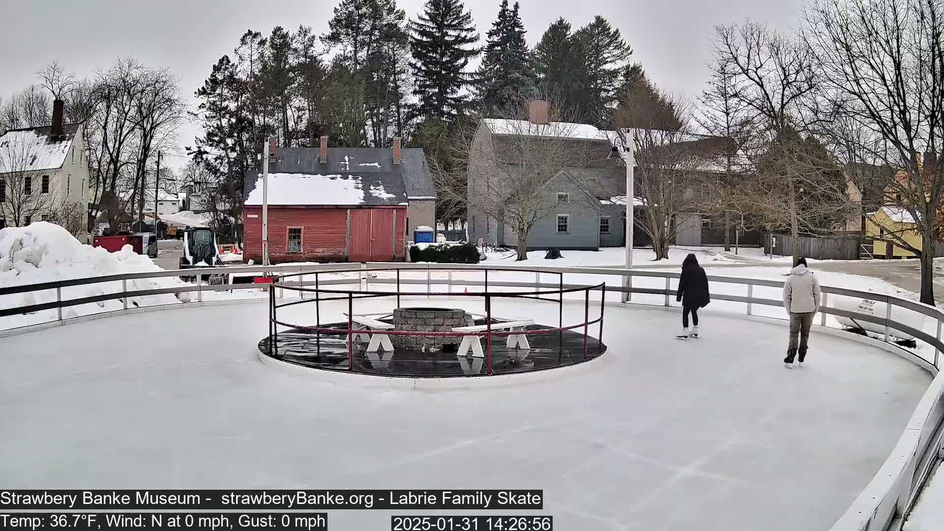 Two people ice skate on an outdoor rink in front of snow-covered buildings on a cloudy day.