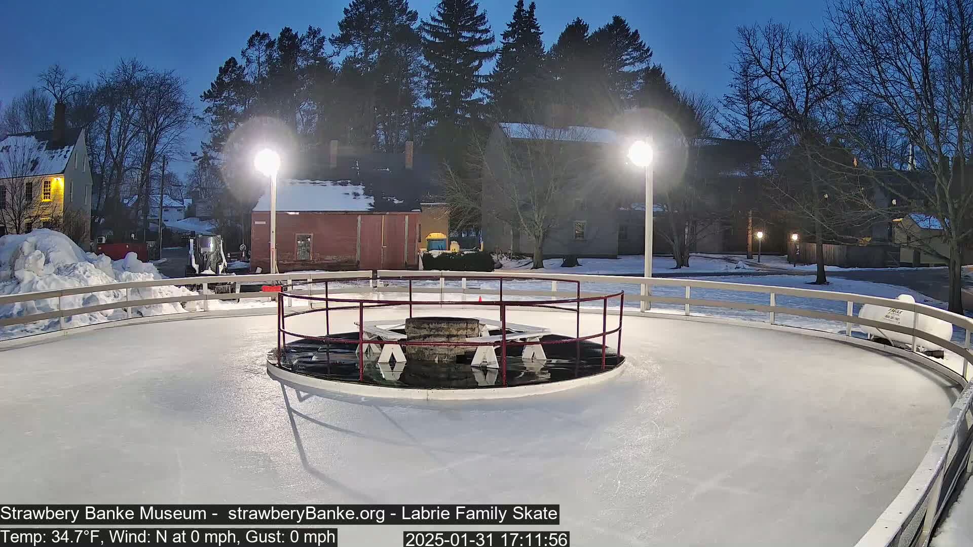 An outdoor ice skating rink, illuminated by lights, is surrounded by a low white railing, with a central fire pit and benches visible under the twilight sky.