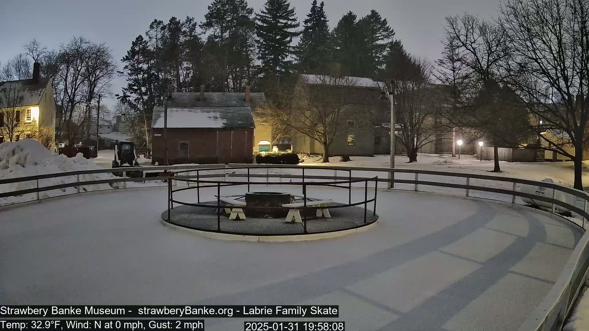A snow-covered outdoor skating rink with a fire pit in the center is surrounded by buildings and trees under a twilight sky.