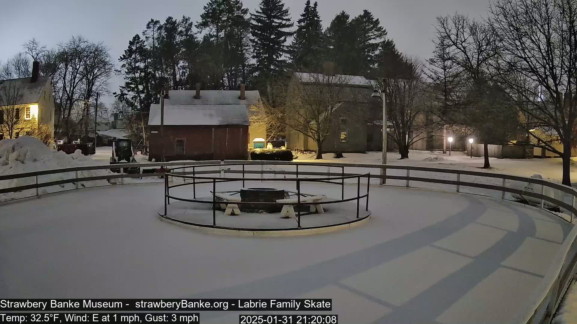 A snow-covered outdoor ice skating rink with a central fire pit is surrounded by a wooden fence and several buildings under a dark, night sky.