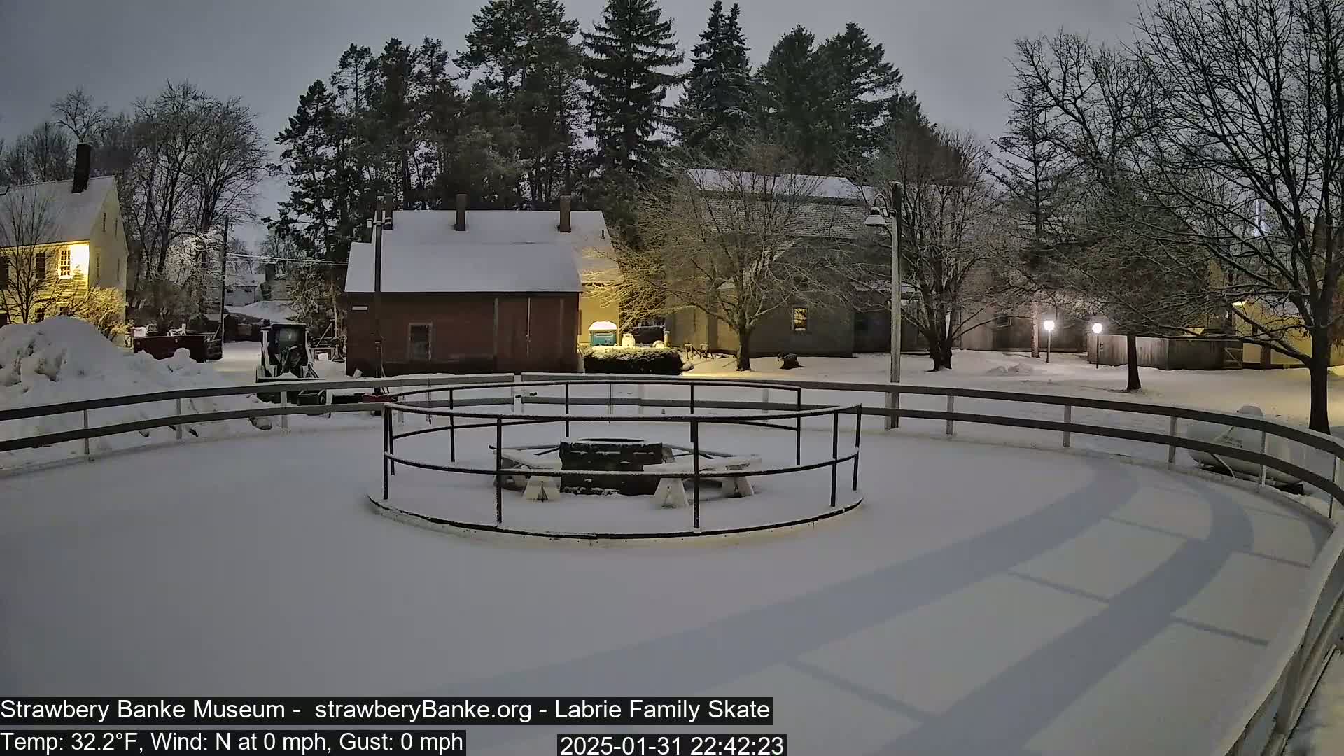 A snow-covered outdoor skating rink with a central fire pit and surrounding buildings under a dark, twilight sky.