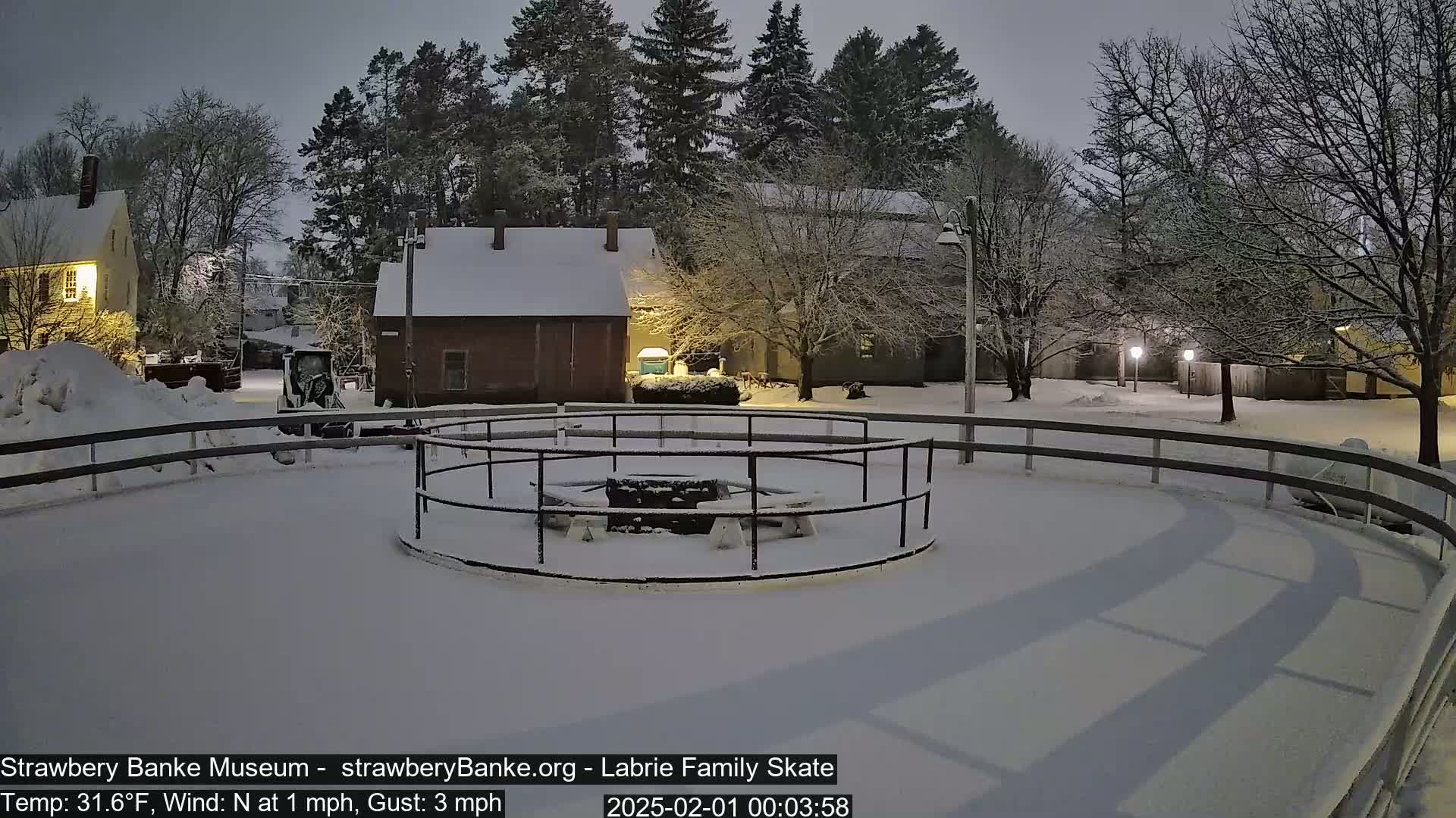 A snow-covered outdoor skating rink with a circular center feature is surrounded by snow-covered buildings and trees under a dark, nighttime sky.