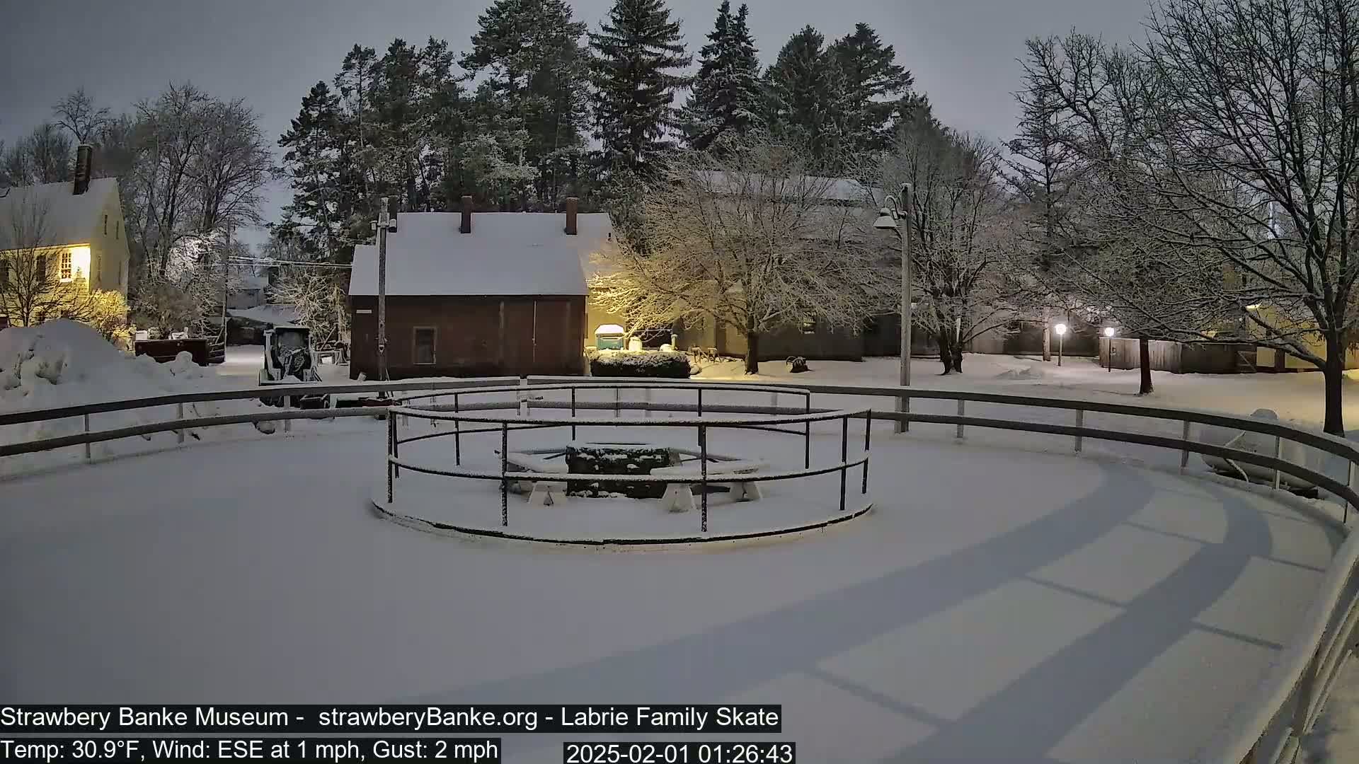 A snow-covered outdoor skating rink with surrounding buildings and trees under a nighttime snowfall.