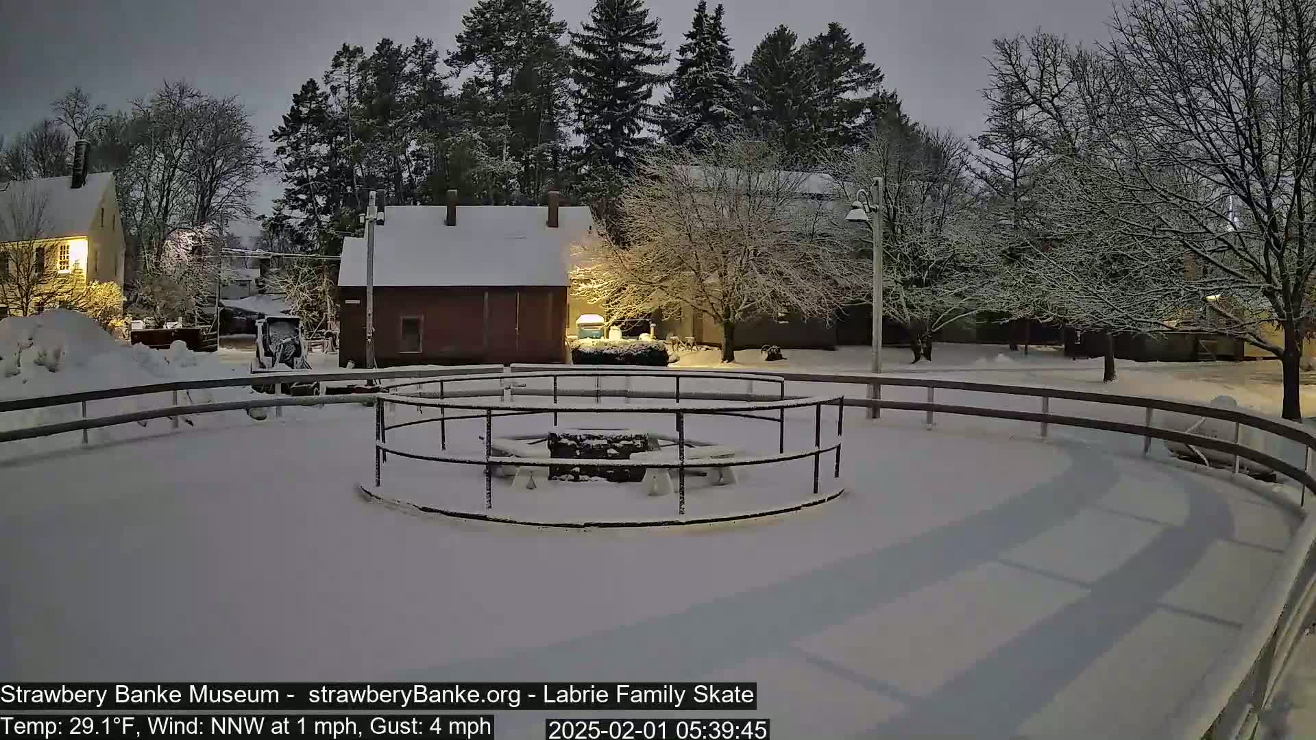 A snow-covered outdoor skating rink is surrounded by snow-covered buildings and trees under a twilight sky during a light snowfall.