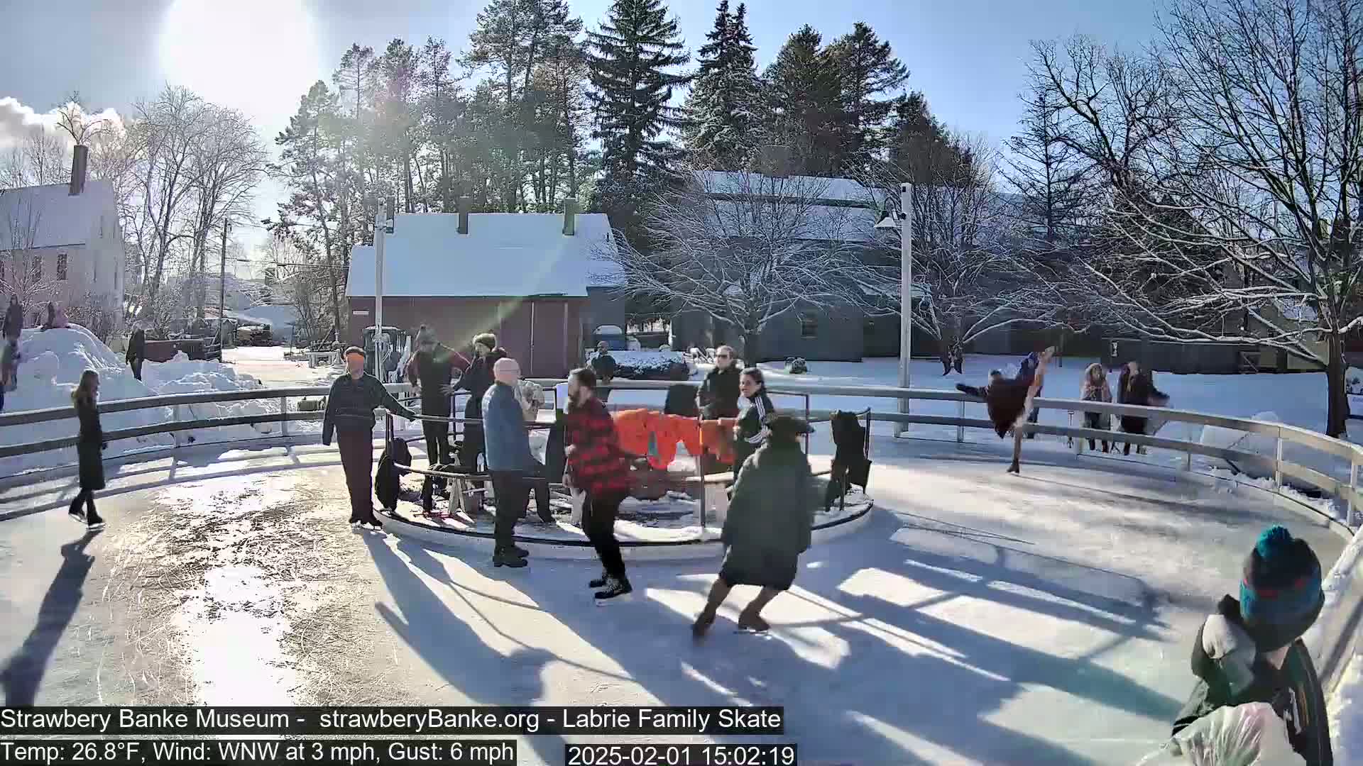 People ice skate on an outdoor rink on a sunny day with a temperature of 26.8°F and a wind from the west-northwest at 3 mph, gusting to 6 mph.