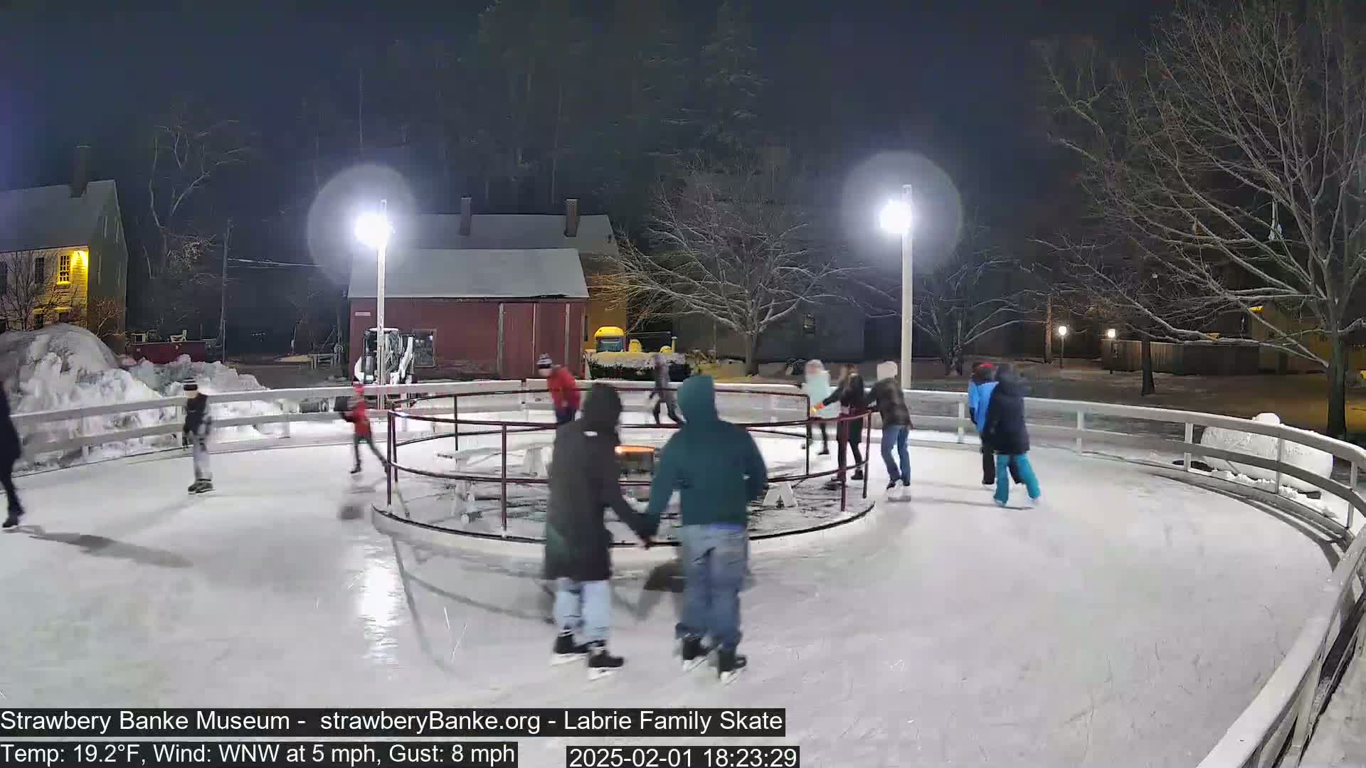 Several people ice skate at night on an outdoor rink illuminated by lights, with snow on the ground and a light wind.
