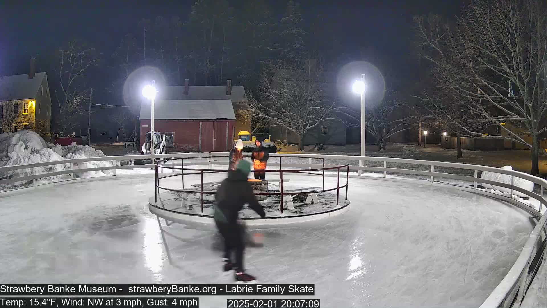 A person ice skates on an outdoor rink at night under light snow, with a small fire pit and several people nearby.