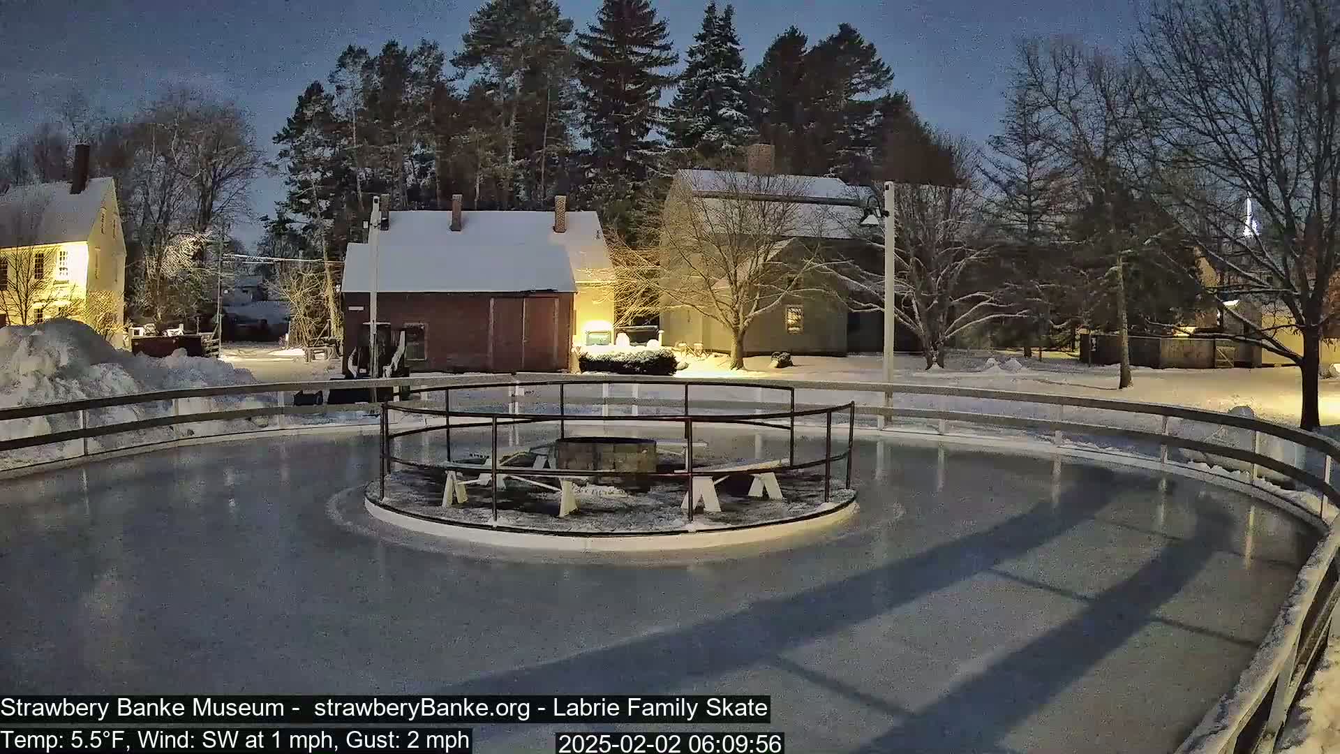 A snow-covered outdoor ice rink, lit at night, is surrounded by snow-covered buildings and trees under a dark sky.