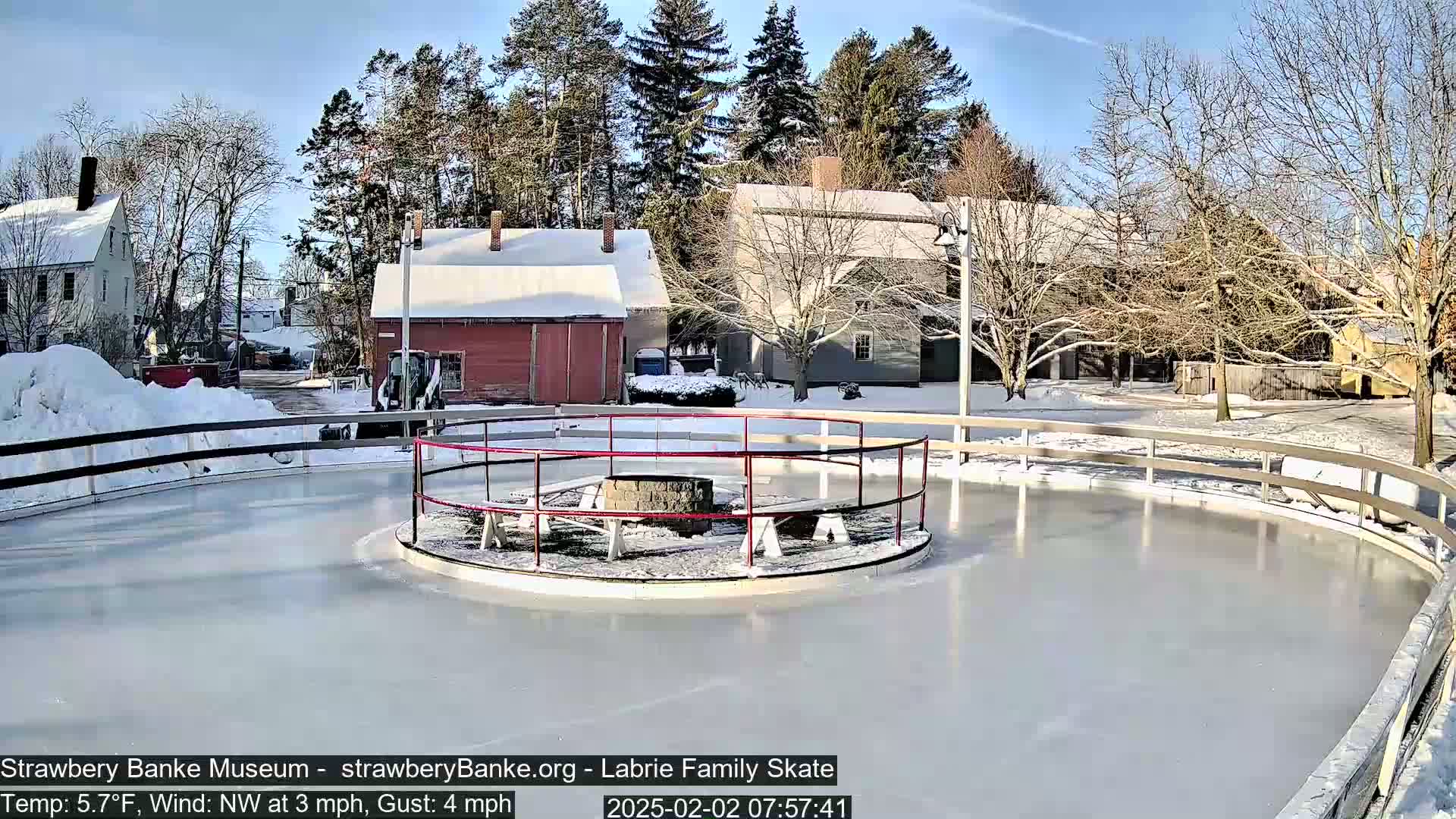 A snow-covered outdoor ice skating rink with a central fire pit, surrounded by several buildings under a sunny sky.