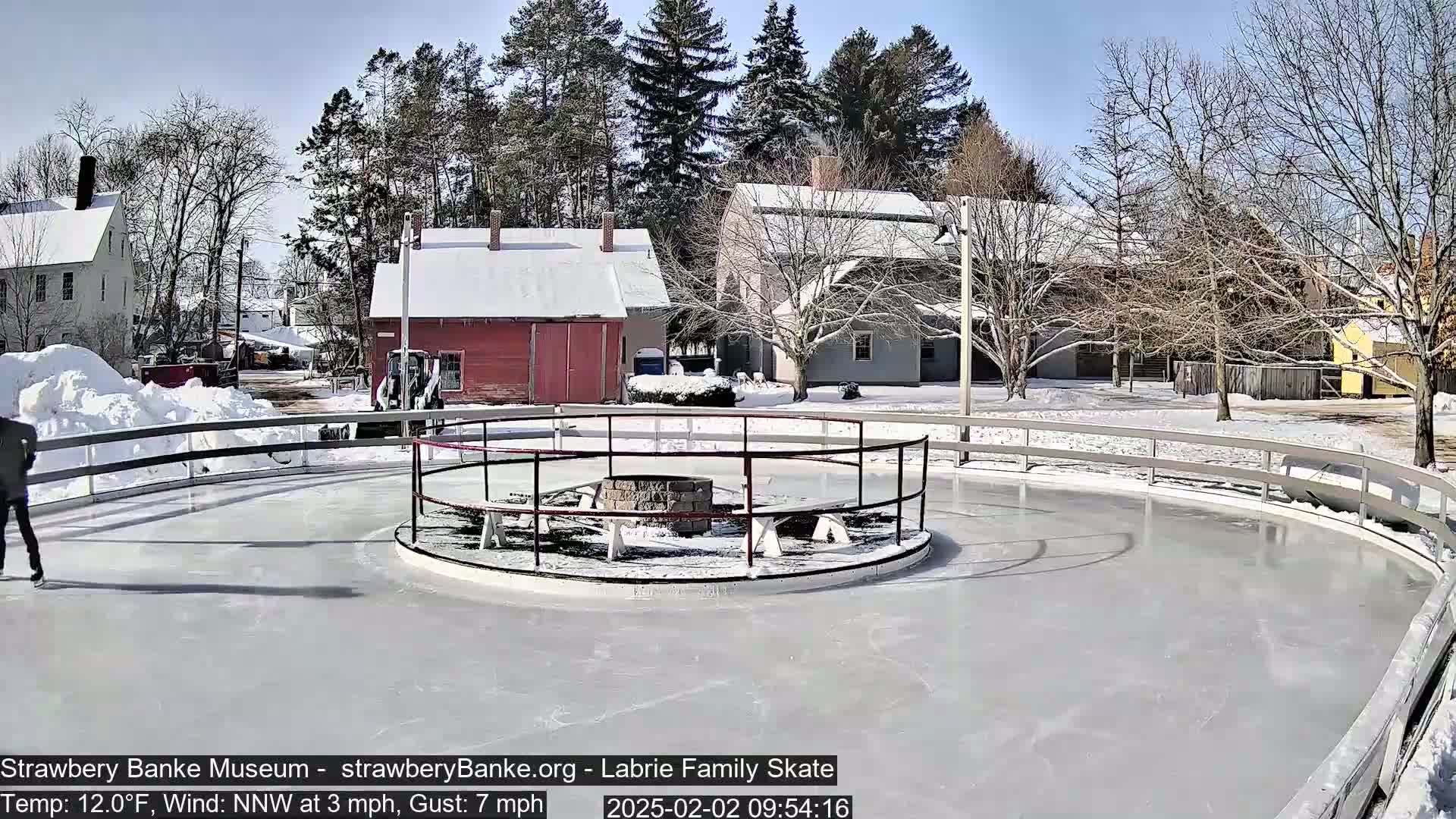 A person ice skates on an outdoor rink surrounded by snow-covered buildings and bare trees under a sunny sky.