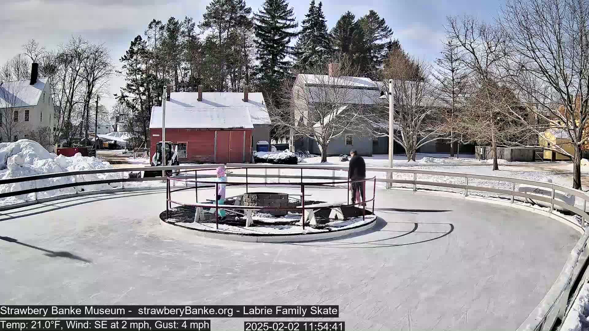 A sunny outdoor ice rink, surrounded by snow-covered buildings and bare trees, features several people ice skating.