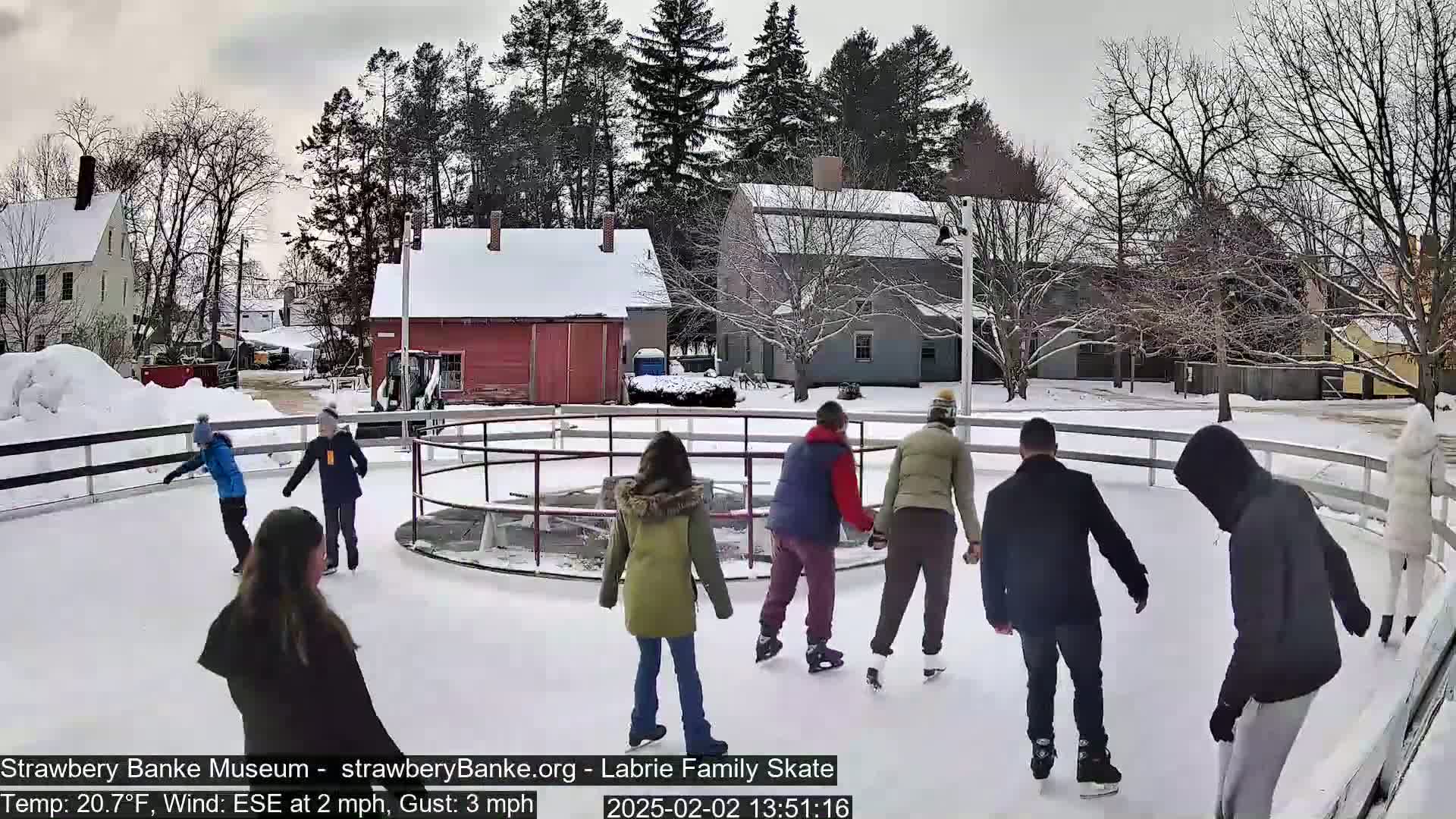 Several people ice skate on an outdoor rink surrounded by a snow-covered fence and buildings under a cloudy sky.