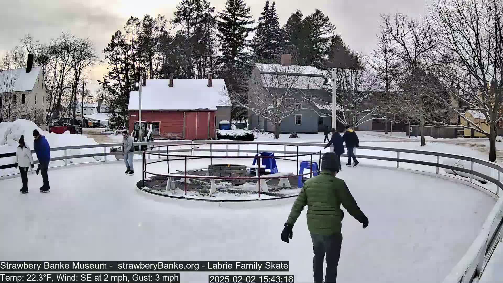 Several people ice skate on an outdoor rink surrounded by snow-covered buildings and trees under a cloudy sky.