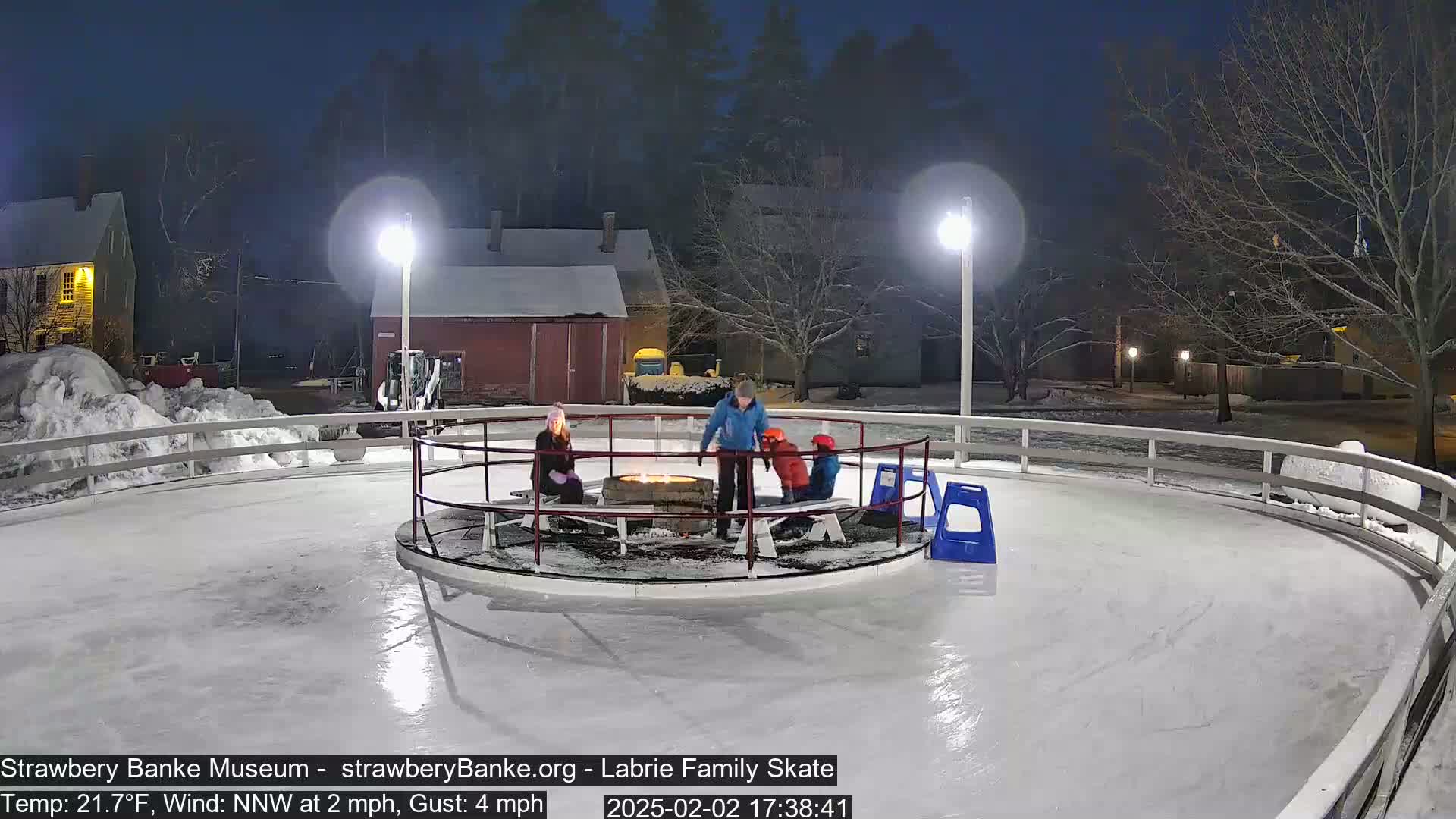 A nighttime outdoor ice skating rink with a small fire pit in the center, surrounded by several people, under lightly snowy conditions.