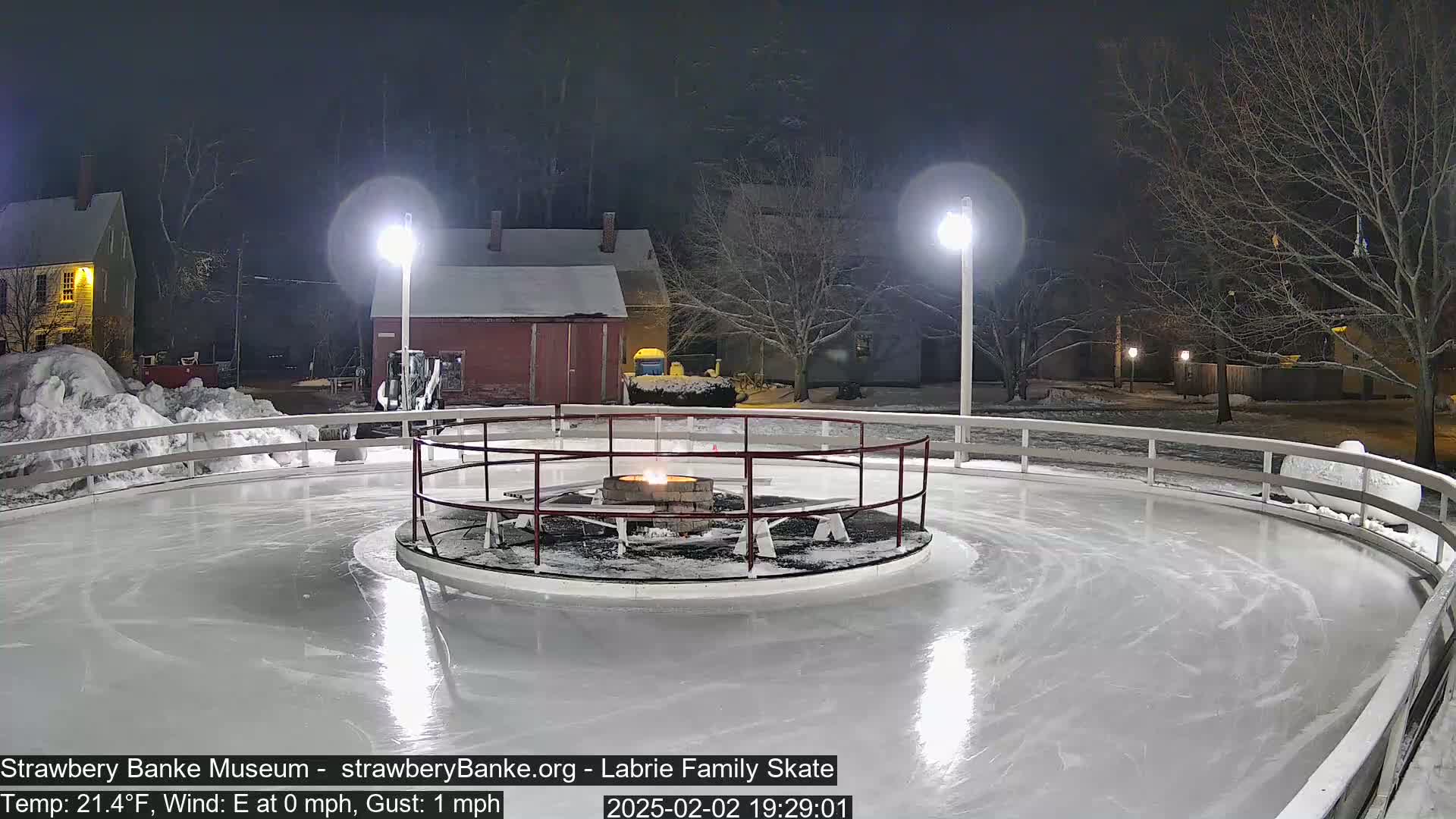 An outdoor ice skating rink is lit at night by two tall lamps, with a fire pit in the center and snow on the ground.