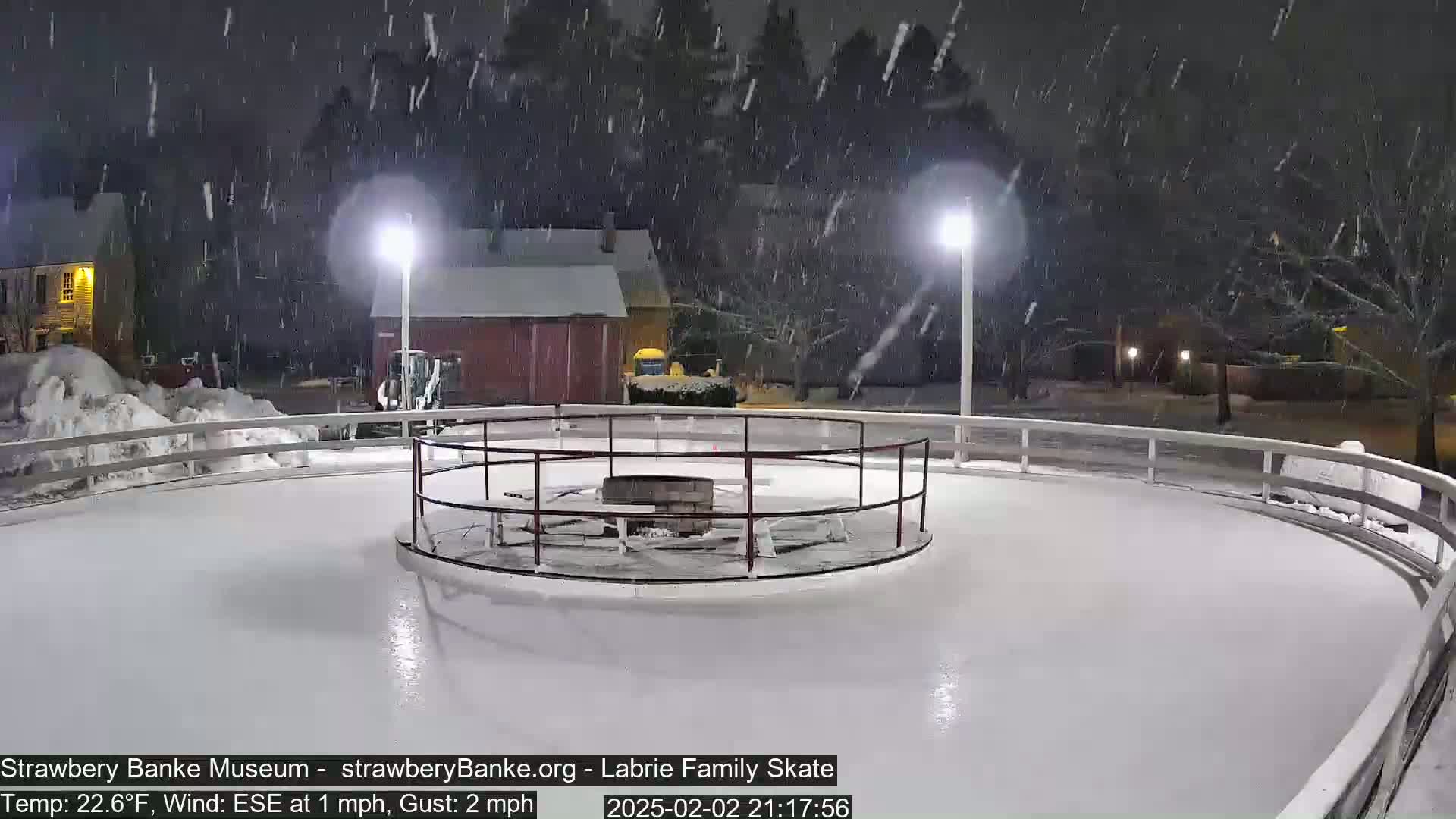 A snow-covered outdoor ice skating rink is illuminated at night under falling snow.