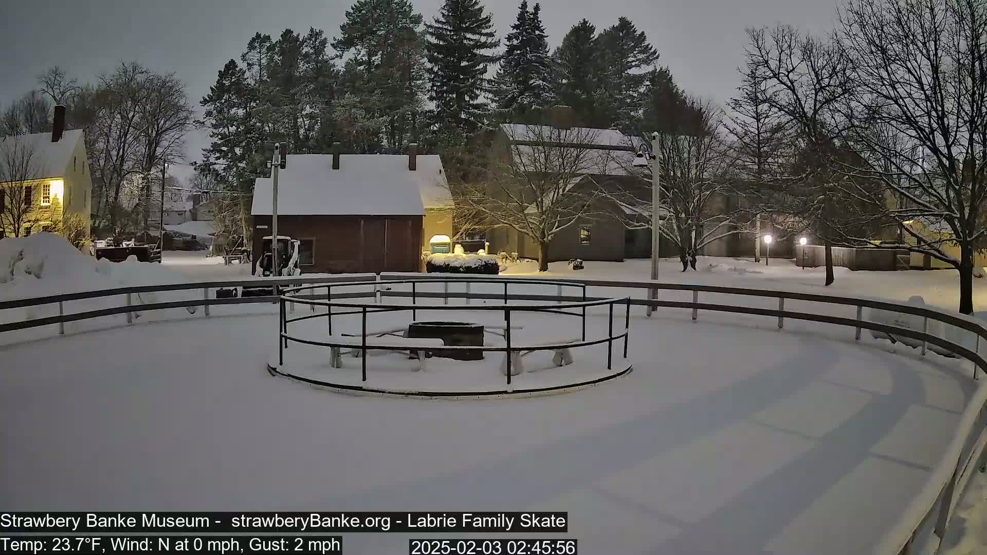 A snow-covered skating rink with a fire pit in the center is surrounded by snow-covered buildings and trees under a dark, snowy night sky.