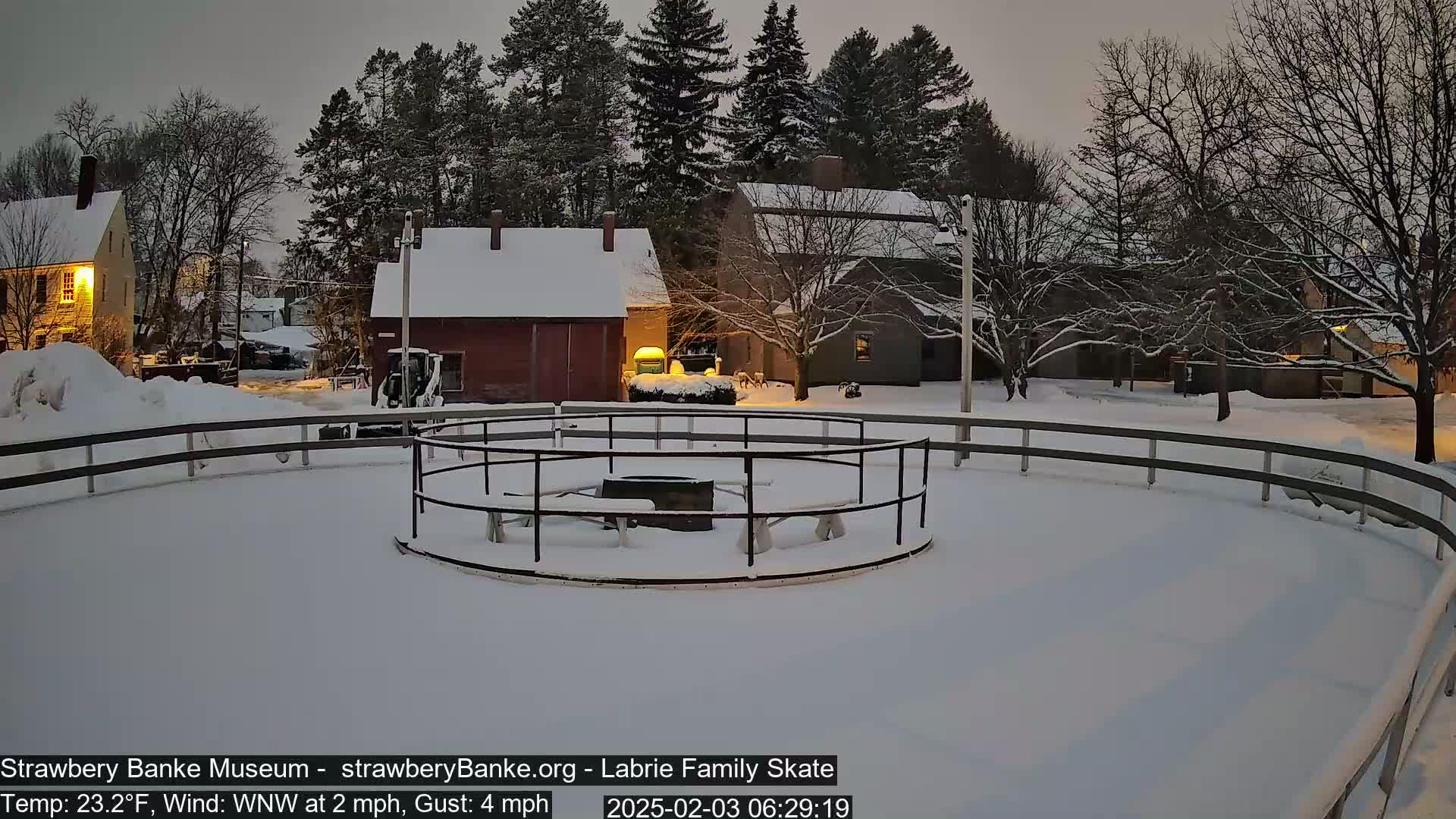 A snow-covered outdoor skating rink with a central fire pit and surrounding buildings under a twilight sky.