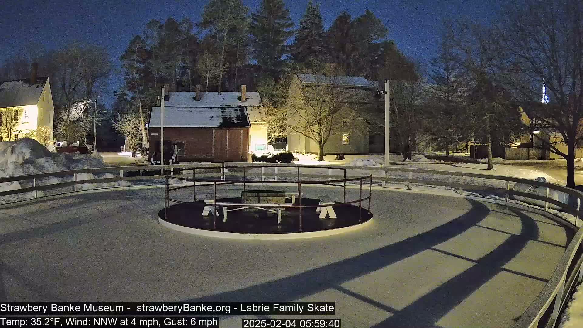 A nighttime view of an outdoor ice skating rink enclosed by a low fence, surrounded by snow-covered buildings and trees under a dark sky.