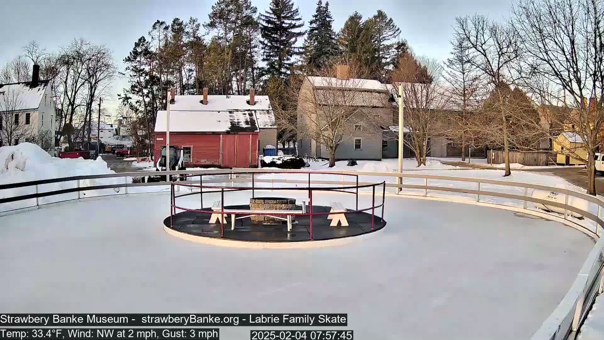 A snow-covered outdoor ice skating rink with a central fire pit and surrounding buildings is visible under a mostly clear sky.