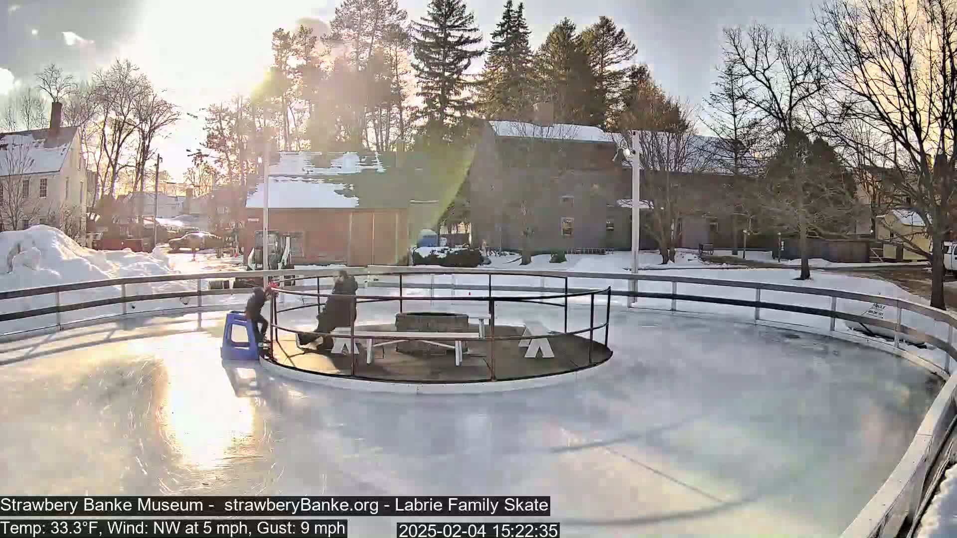 An outdoor ice skating rink, partially sunny with a northwest wind, features two people near a central fire pit and seating area.