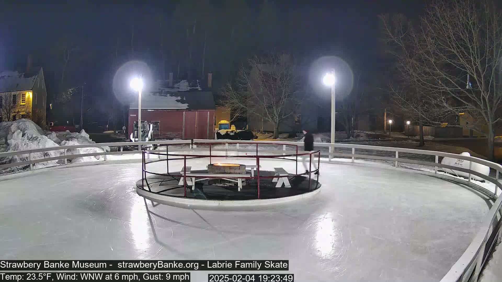 A nighttime outdoor ice skating rink with a fire pit in the center, illuminated by lights, surrounded by buildings and bare trees.