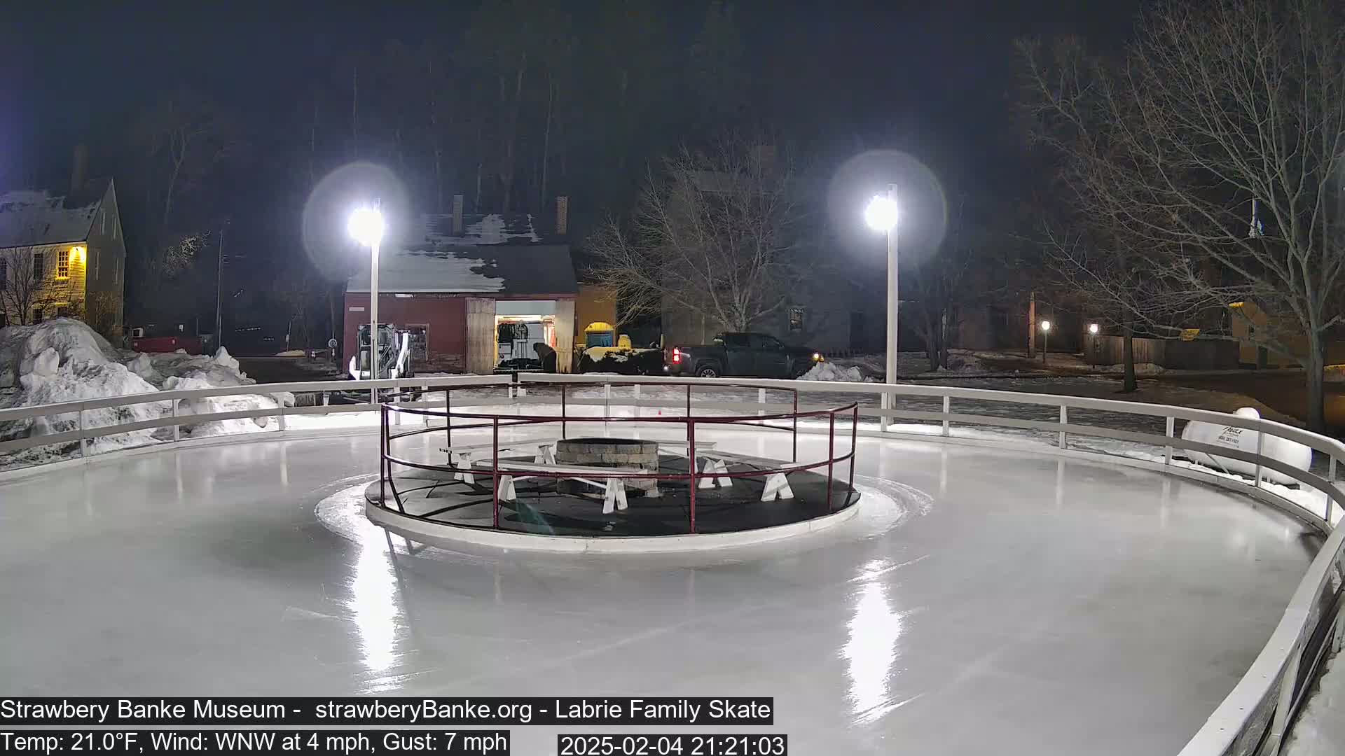 An outdoor ice skating rink, illuminated at night by two lampposts, is mostly empty under a dark, clear sky; a fire pit sits in the center of the rink.