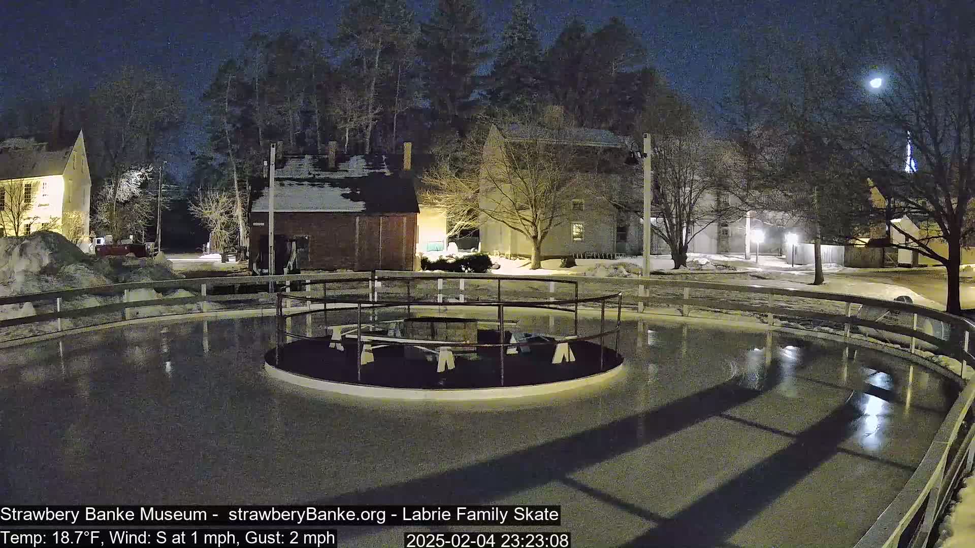 An outdoor ice skating rink, illuminated at night under a mostly clear sky, is surrounded by snow-covered ground and buildings.