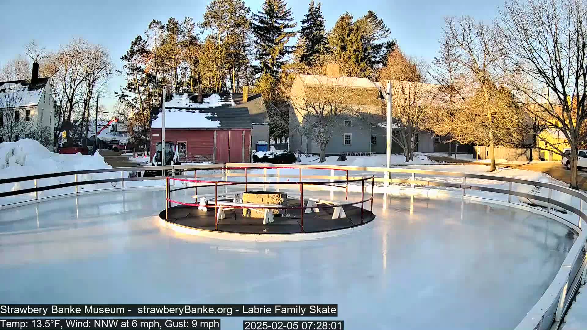 A circular outdoor ice skating rink with a central fire pit and benches, surrounded by snow-covered buildings and trees under a clear, sunny sky.