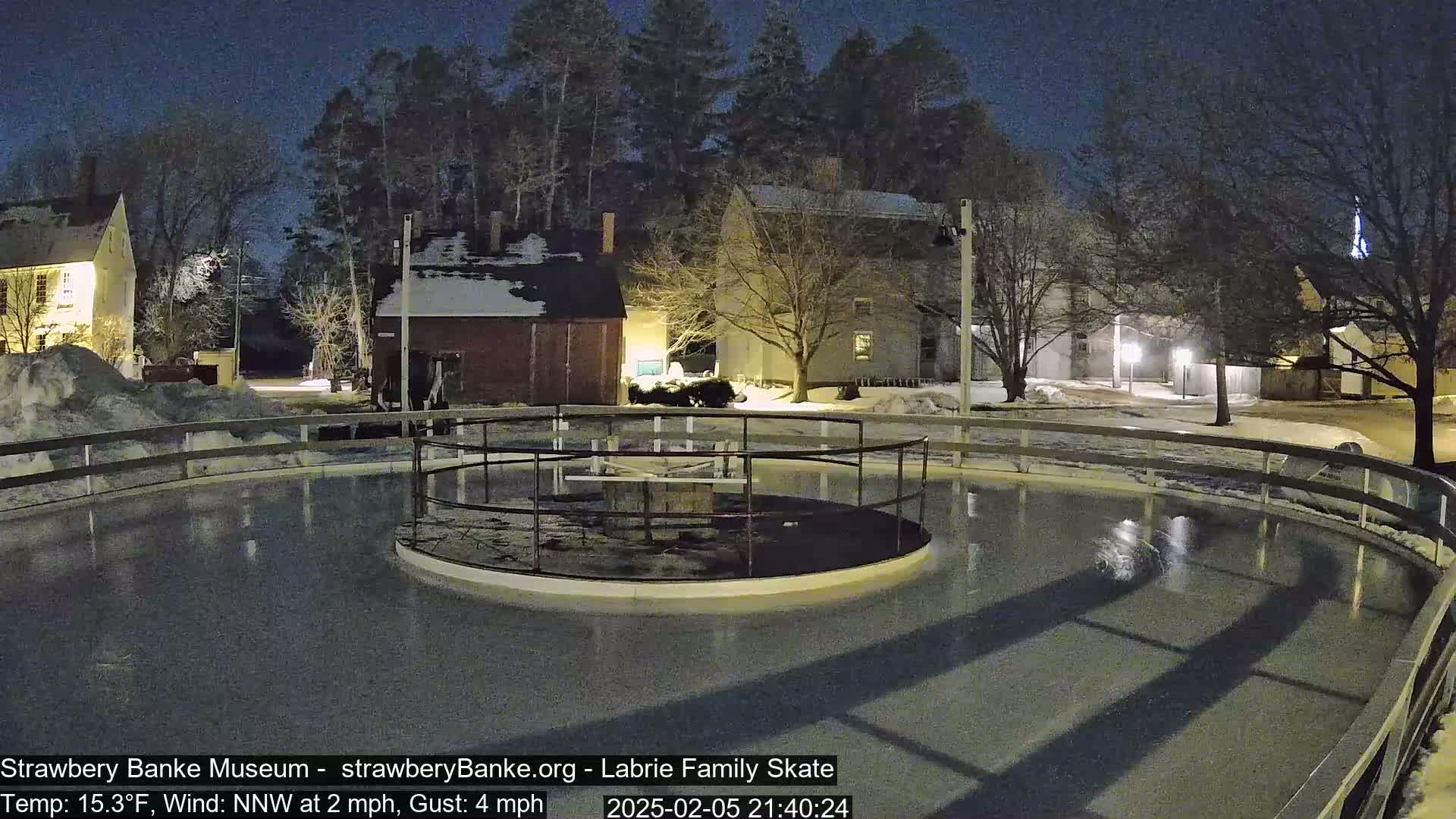 An outdoor ice rink, surrounded by snow-covered buildings and trees, is illuminated at night under a dark sky.