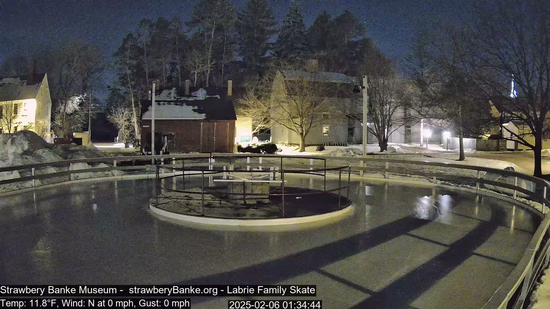 An empty outdoor ice rink is illuminated at night, surrounded by snow-covered ground and buildings under a clear, dark sky.