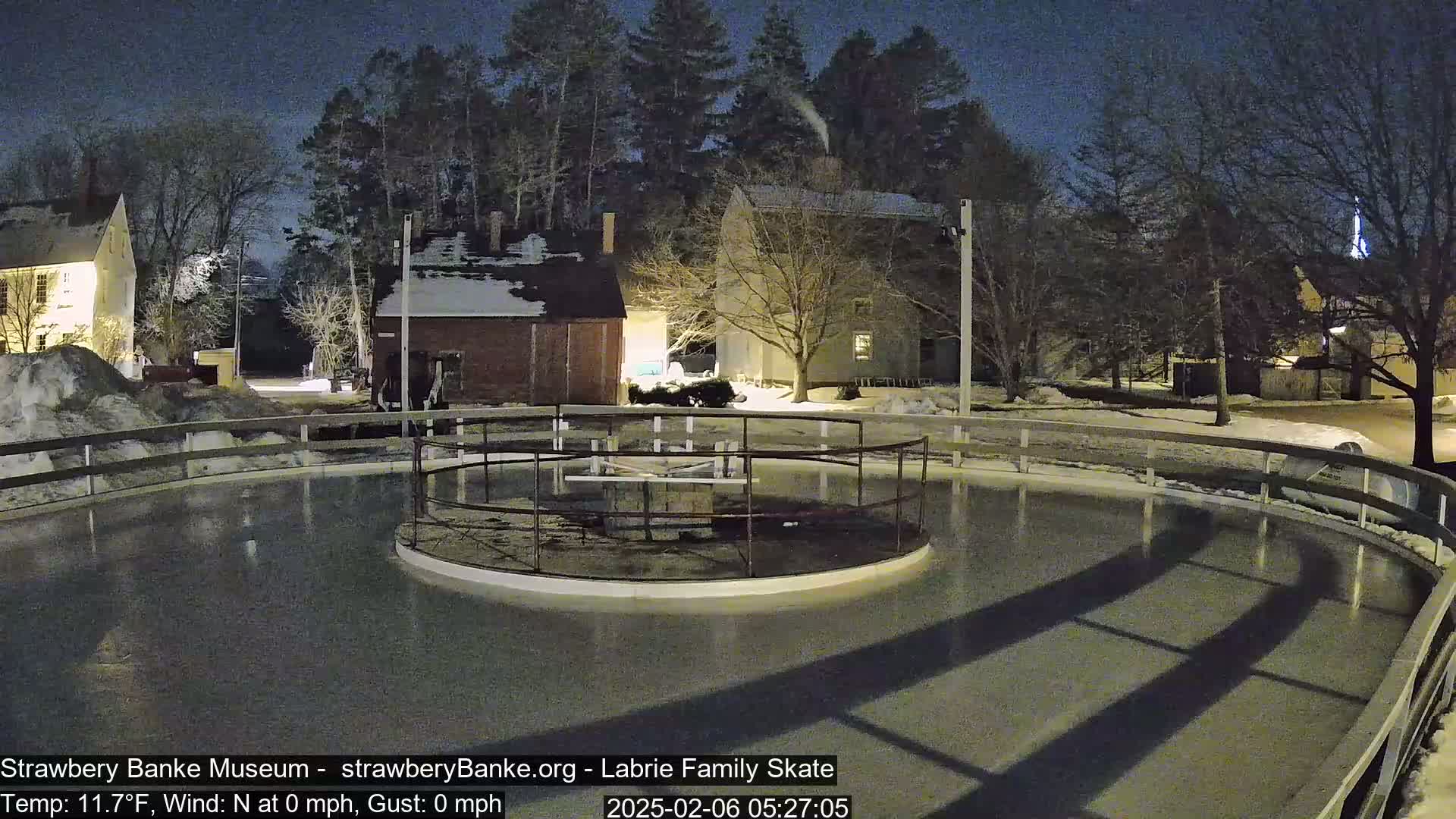 A nighttime view of an outdoor ice rink surrounded by snow-covered ground and historic-looking buildings under a dark sky.