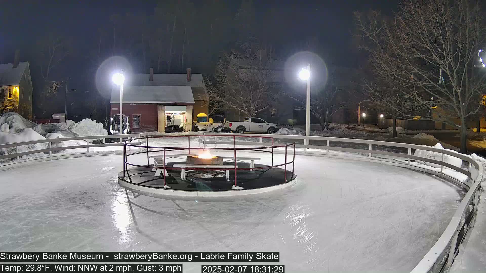 A nighttime view of an outdoor ice skating rink with a fire pit in the center, surrounded by snow and lit by lamps.