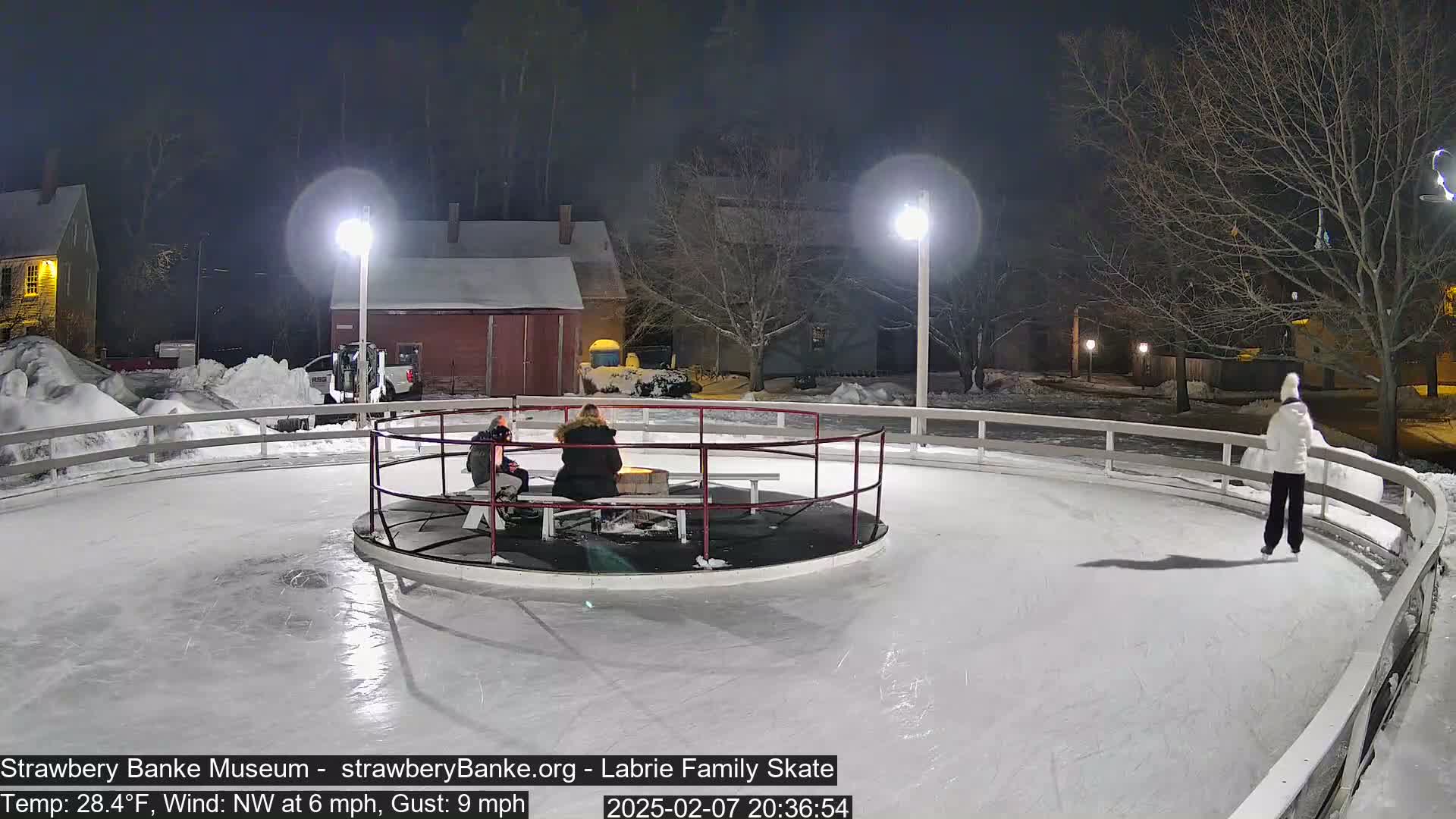 A nighttime scene shows an outdoor ice skating rink with several people ice skating and others sitting near a fire pit, under illuminated lamp posts and surrounded by snow.