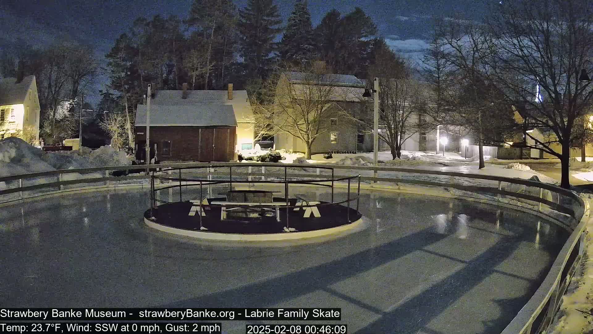 An outdoor ice skating rink, enclosed by a fence, is illuminated at night under a mostly clear sky, with snow on the ground and nearby buildings.